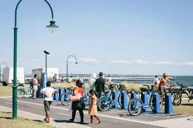 People walking and biking along South Melbourne promenade with blue bike racks under a clear sky.