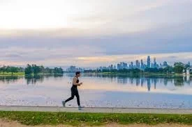A person jogging along Albert Park akeside path with a Melbourne skyline in the background during overcast weather.