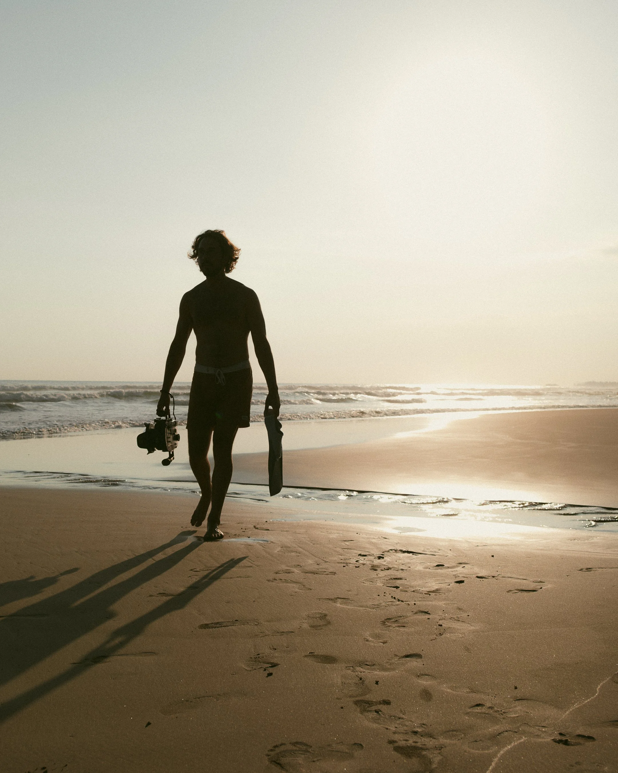 Silhouette d'une personne marchant sur la plage au coucher du soleil, portant du matériel de plongée.