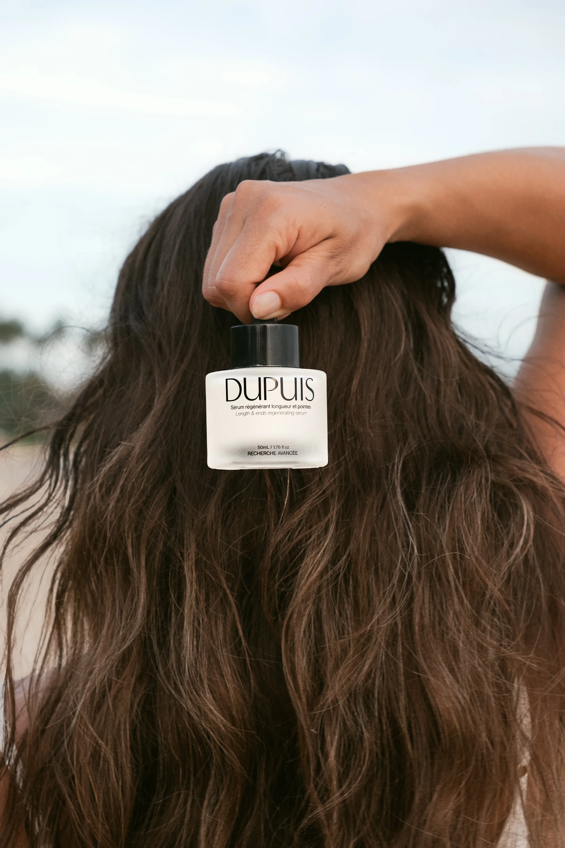 Main tenant un flacon de sérum "DUPUIS" devant des cheveux longs et ondulés.