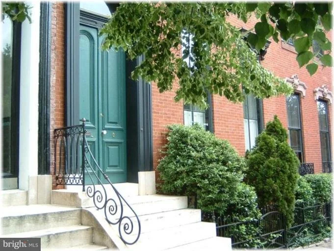 Green front door with black frame, white stairs and black decorative handrail, surrounded by green bushes and trees, red brick building, and windows with white frames.