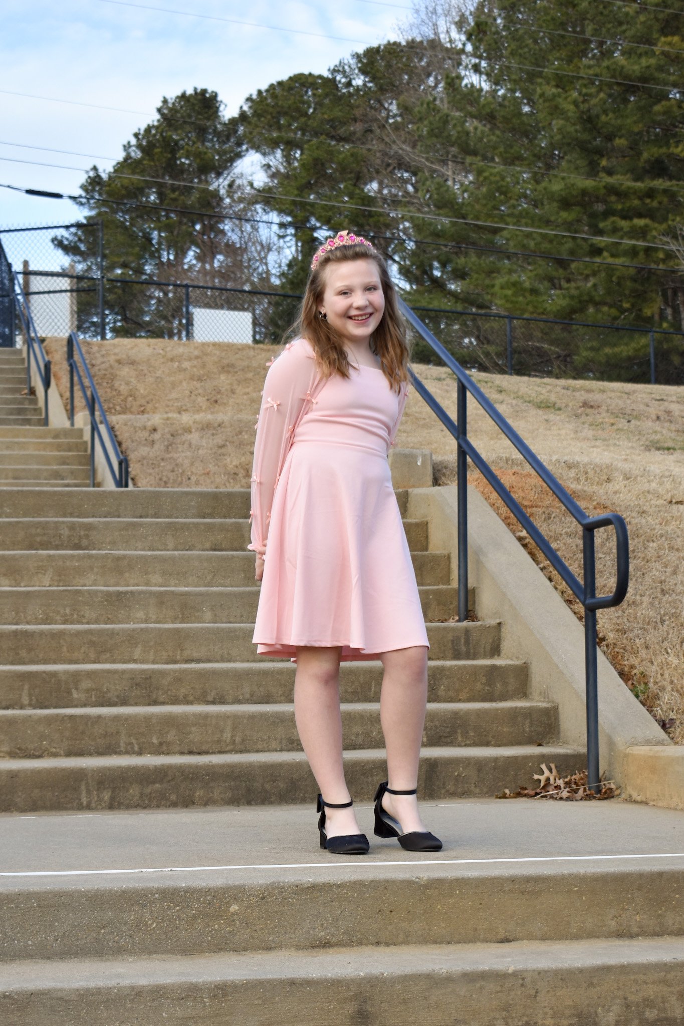 Young girl in a pink dress and black heels standing on outdoor stairs, smiling at the camera.