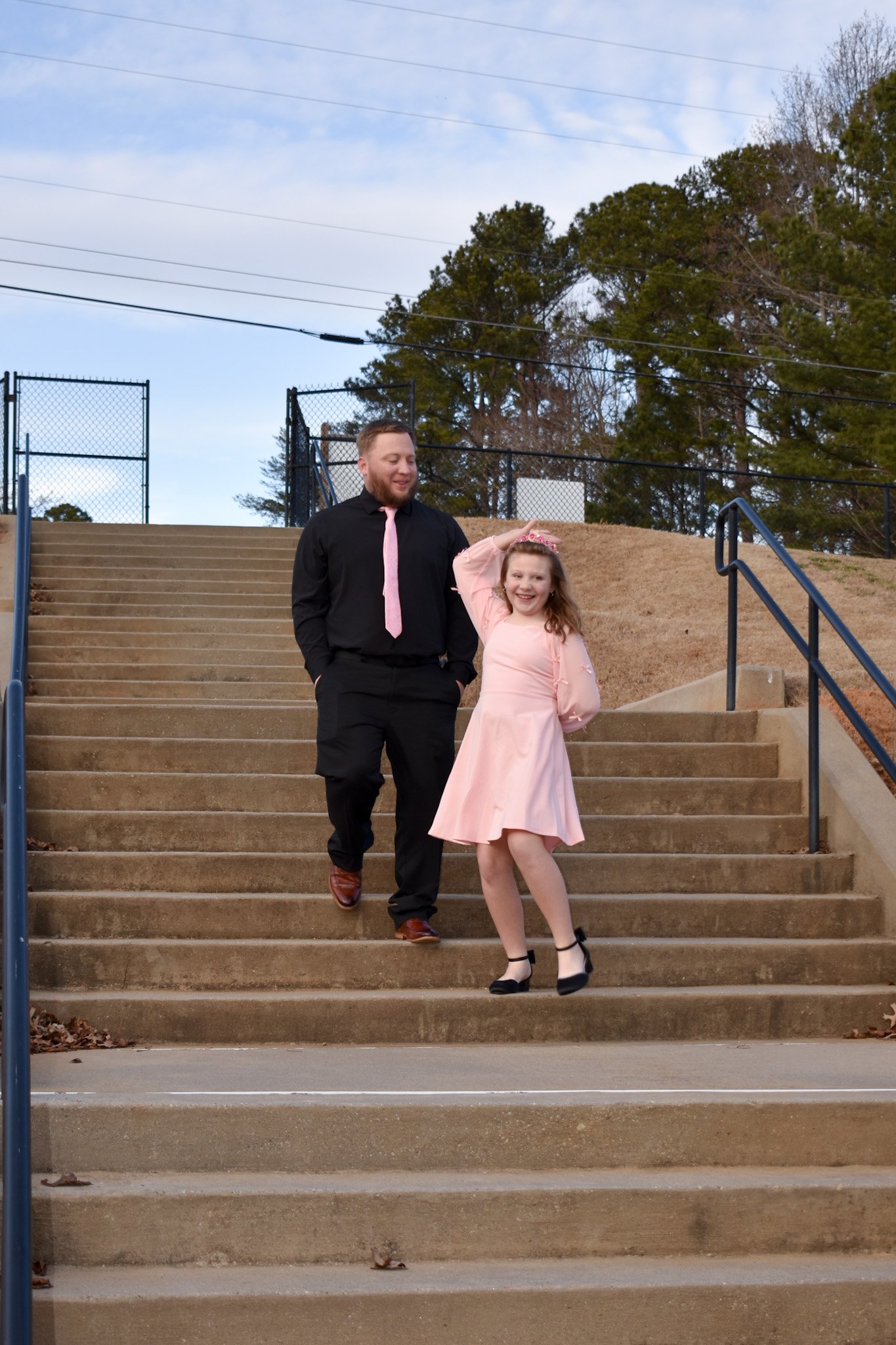 A man and a young girl walking down outdoor concrete stairs. The man is wearing a black shirt, pink tie, and black pants, smiling at the girl. The girl is in a pink dress with black shoes, smiling and posing with her right hand on her head. Trees and