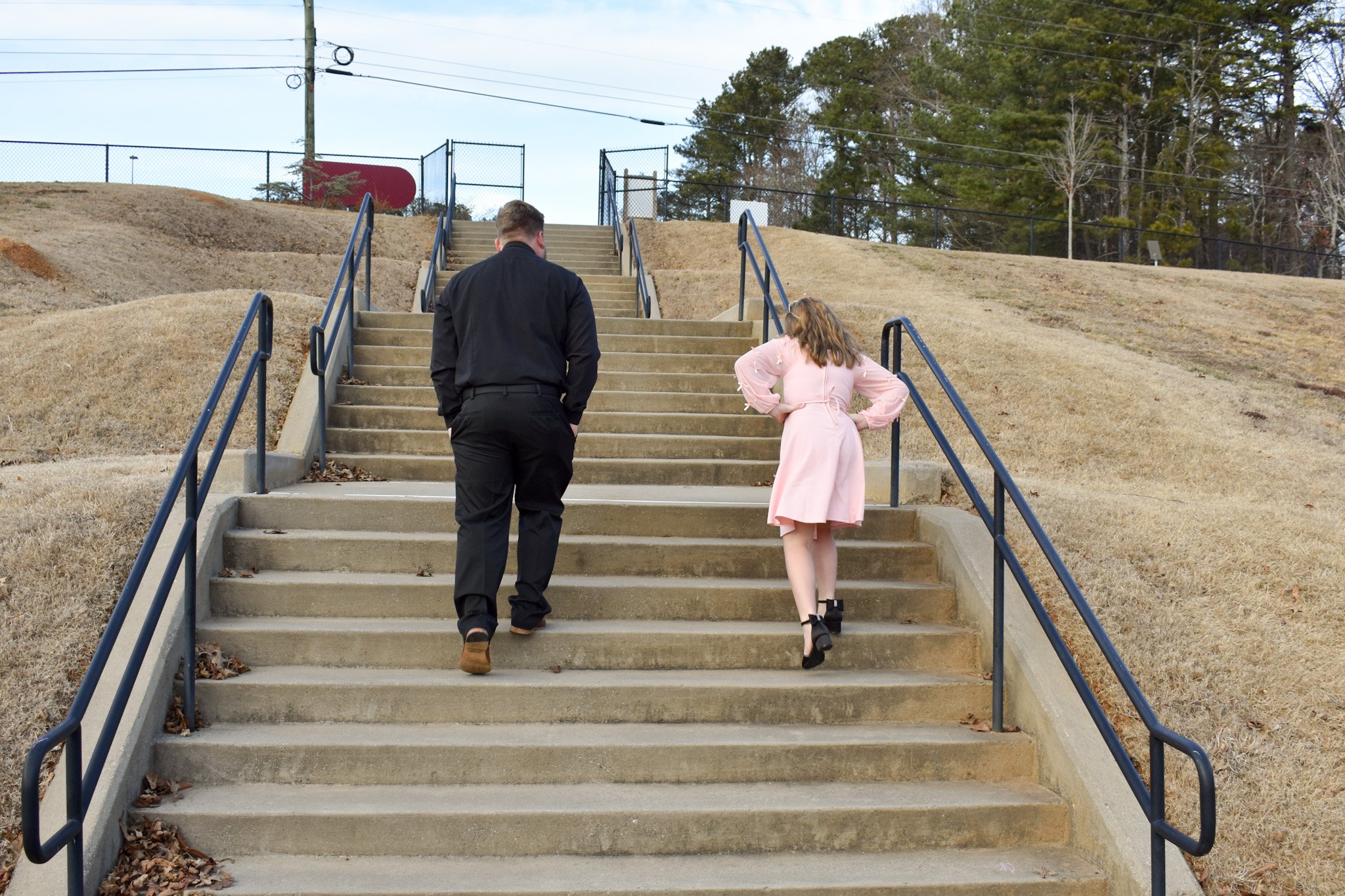 A man and a woman walking up an outdoor staircase with black railings, surrounded by grassy hills and trees, under a cloudy sky.