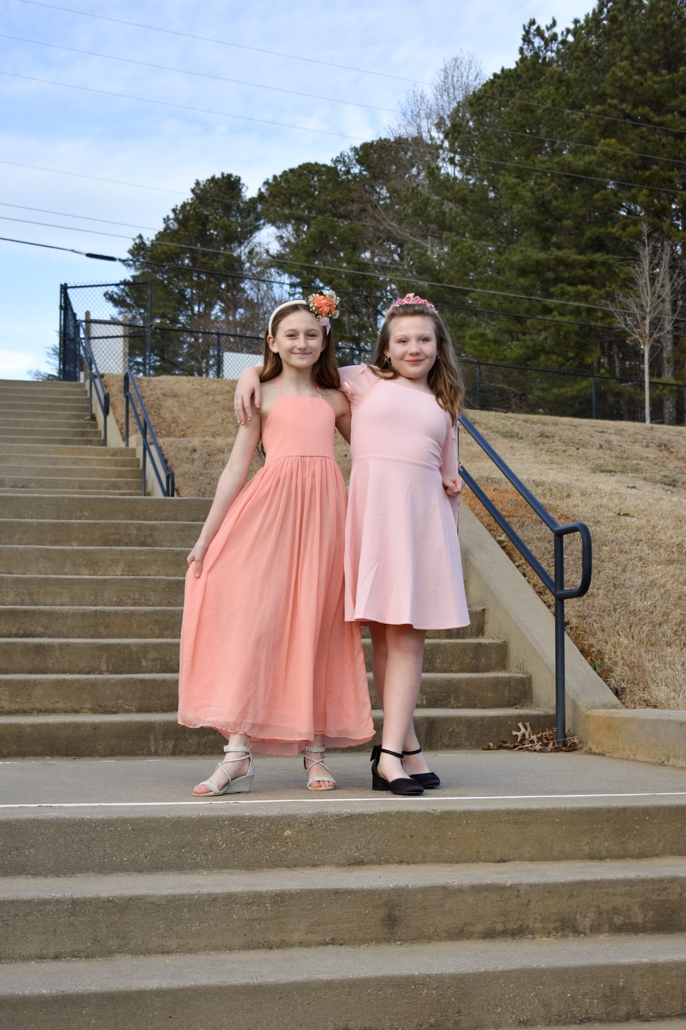 Two young girls in pastel dresses stand close together on outdoor concrete stairs, with one girl's arm around the other's shoulders, smiling at the camera. One girl wears a peach-colored maxi dress with strappy sandals, and the other wears a light pi