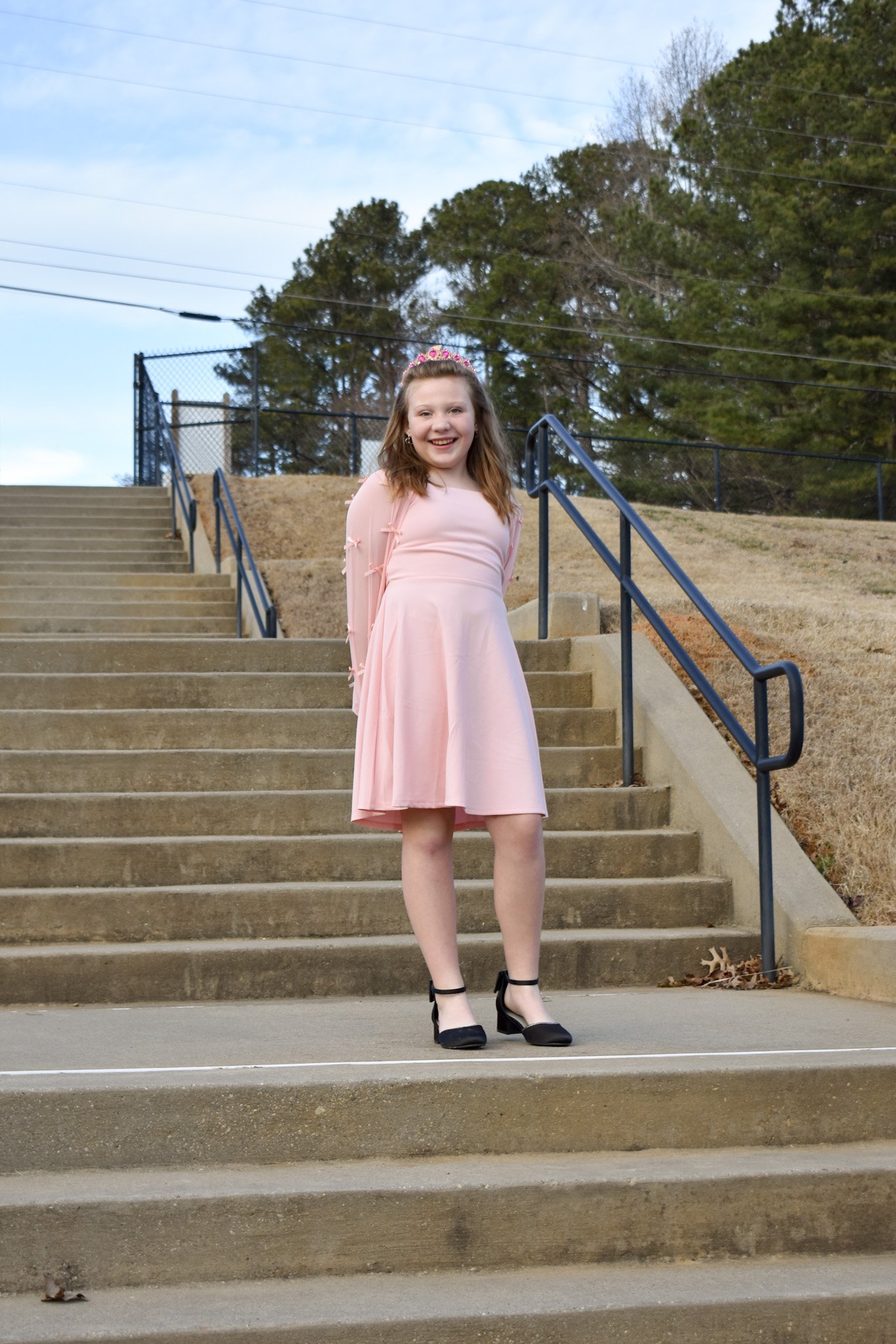 A young girl in a pink dress and black shoes standing on outdoor concrete stairs, smiling at the camera with trees in the background.