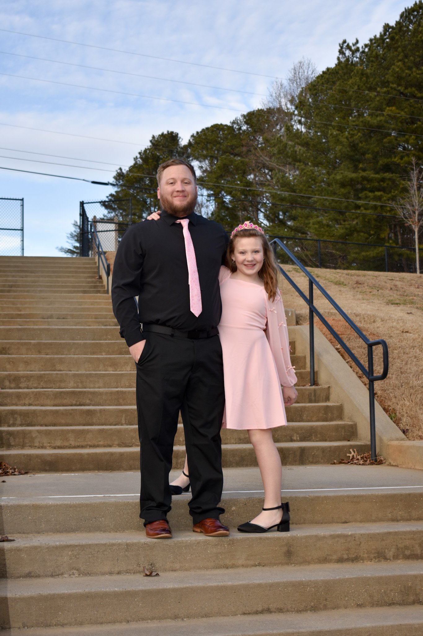 A man and a girl standing together on outdoor stairs, smiling. The man is wearing a black shirt with black pants and a pink tie, while the girl is dressed in a pink dress with a matching tiara.