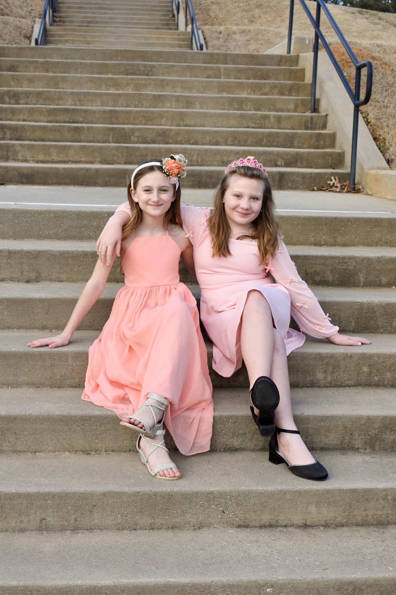 Two young girls sitting on outdoor concrete stairs, wearing pink dresses and tiaras, smiling at the camera.