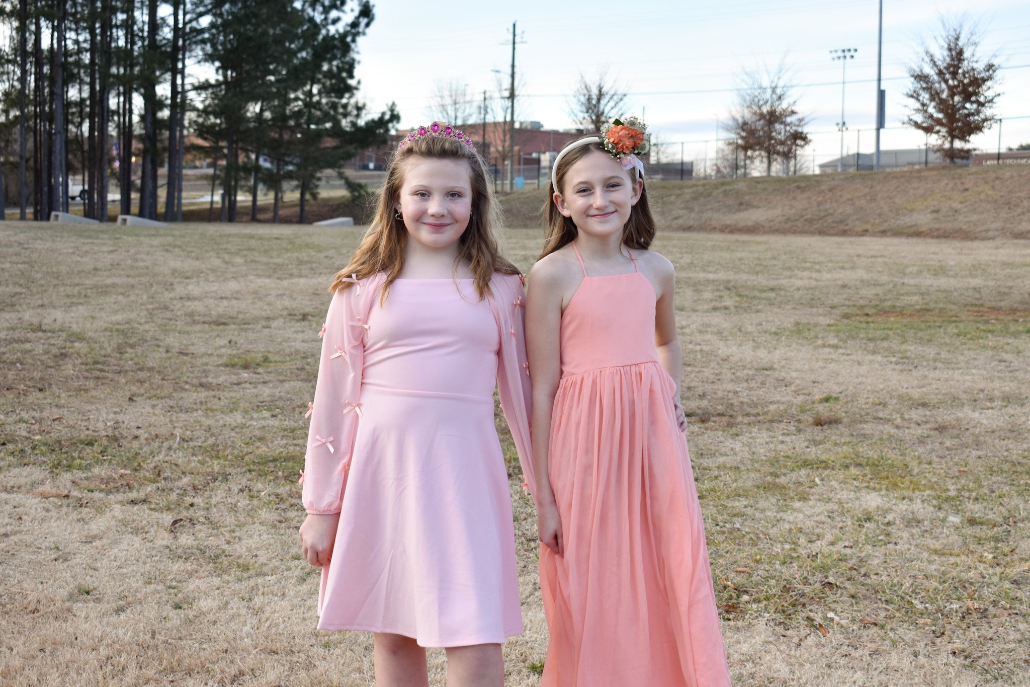 Two young girls in pink dresses standing outdoors on a grassy area, smiling at the camera.