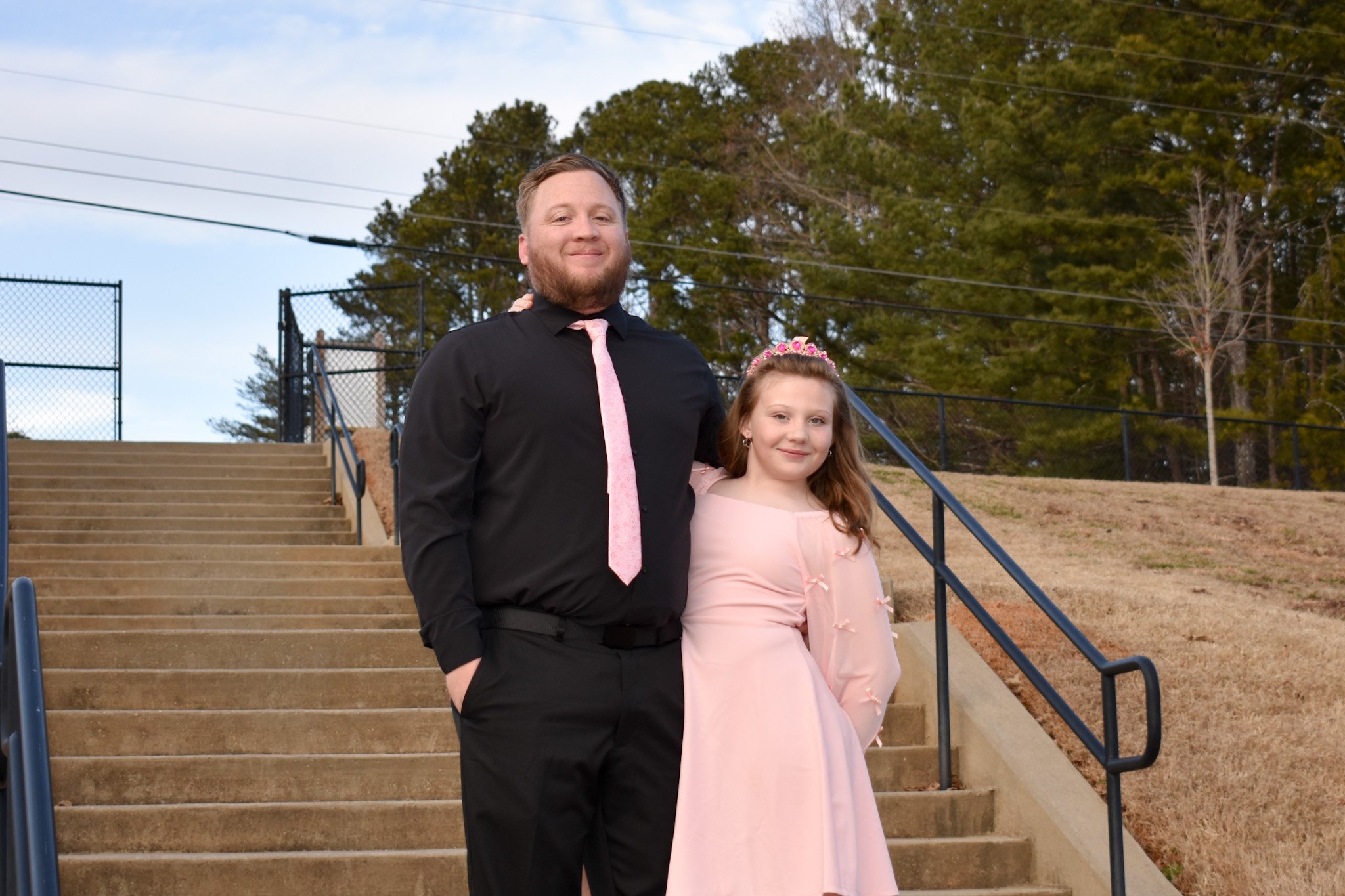 A man and a young girl standing on outdoor stairway, smiling, with trees in the background. The girl is dressed in a light pink dress with small bow accents, wearing a tiara. The man is dressed in black with a pink tie.