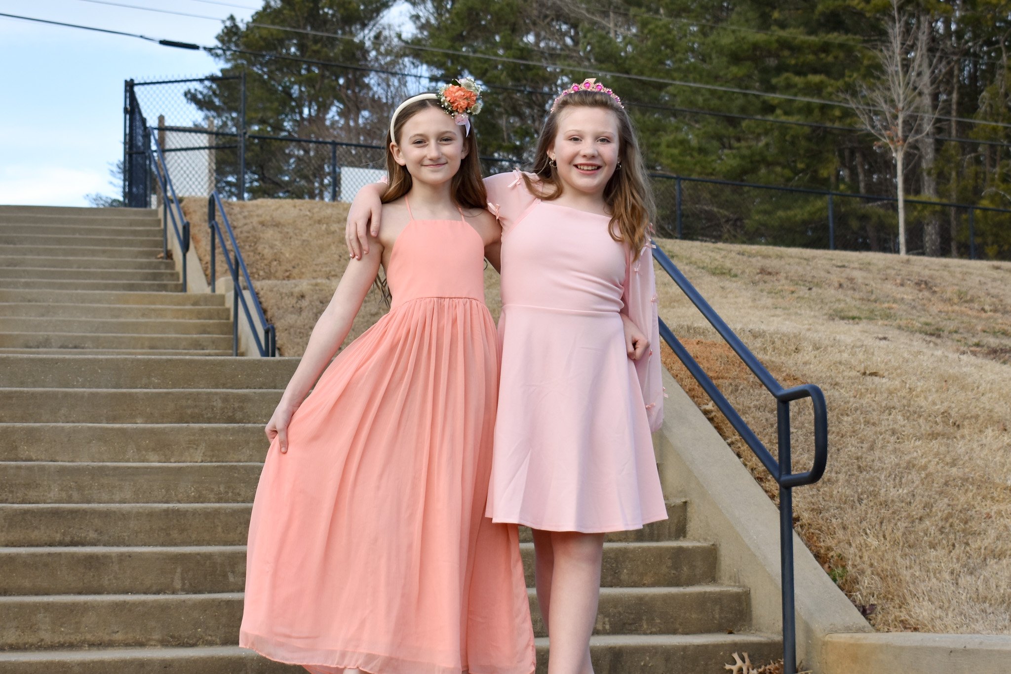 Two young girls in pink dresses and flower crowns smiling and standing side by side on outdoor stairs, with one girl's arm around the other's shoulders.