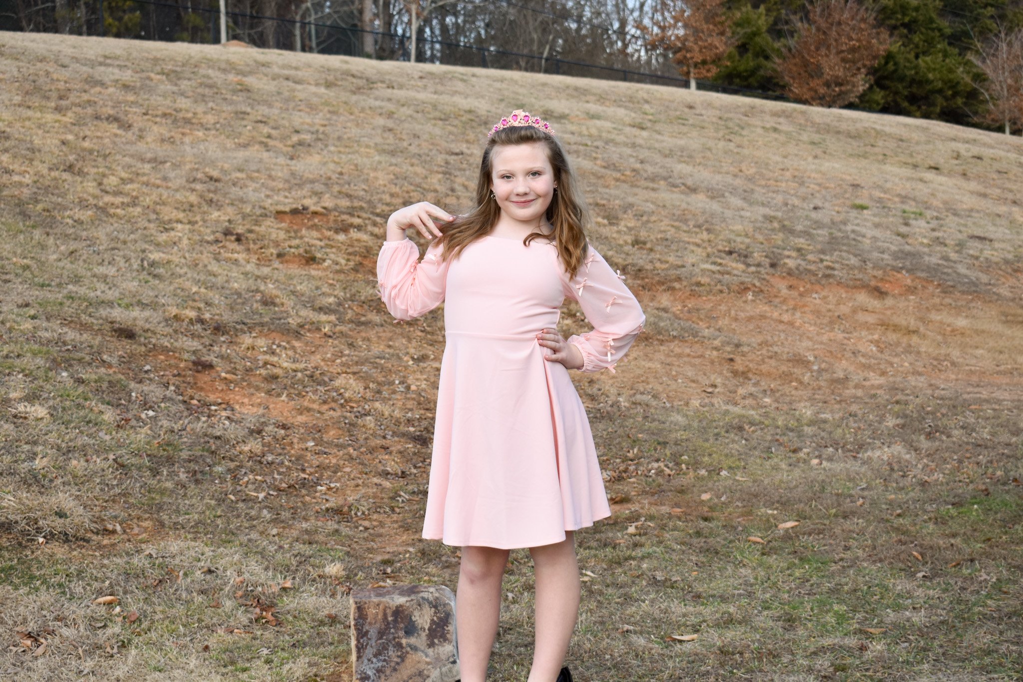 A young girl wearing a pink dress and a pink tiara standing outdoors on a grassy, slightly sloped area with a few trees in the background.