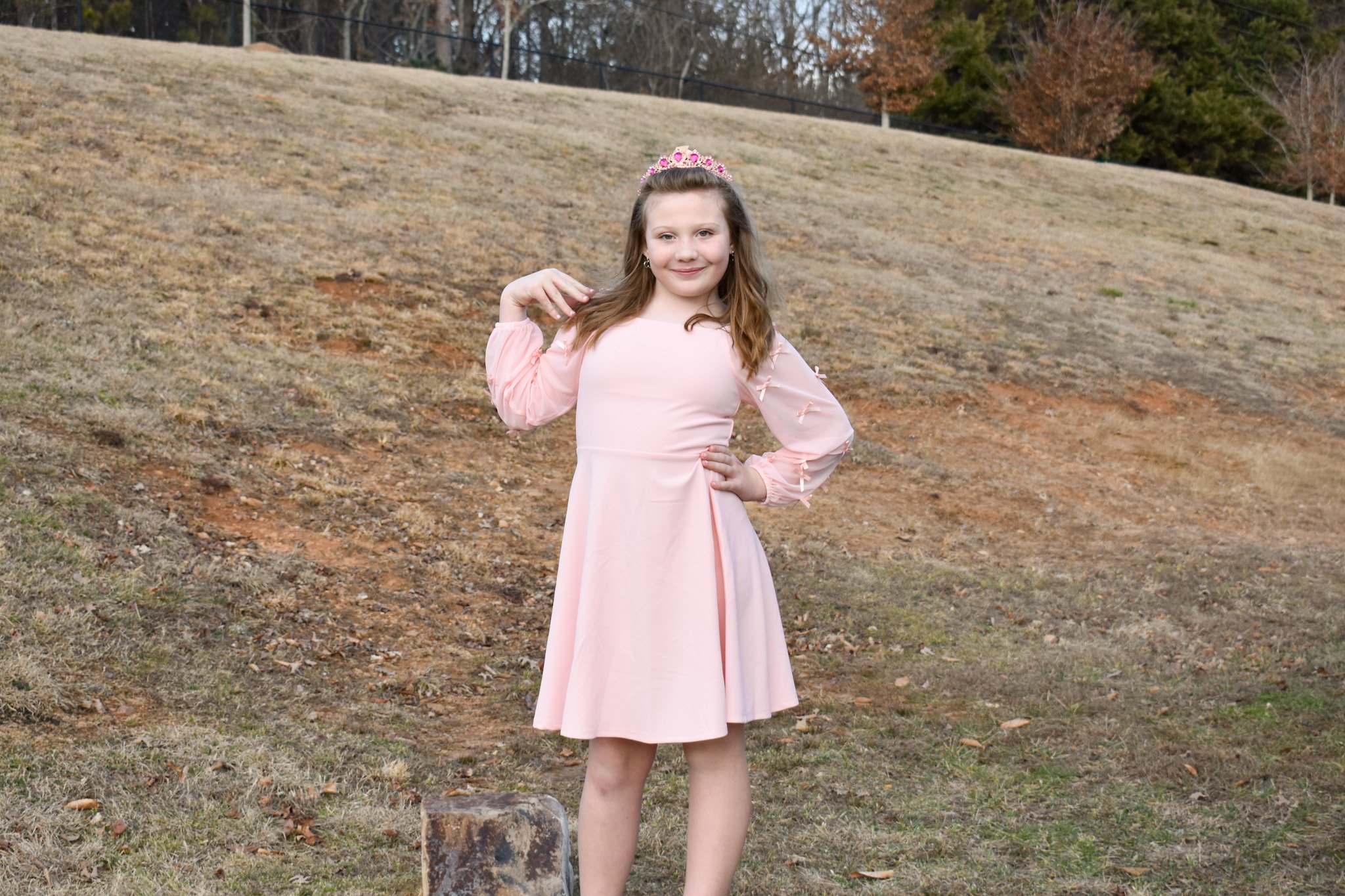 A young girl with long hair wearing a light pink dress and a pink tiara, standing outdoors on a grassy area with trees in the background.