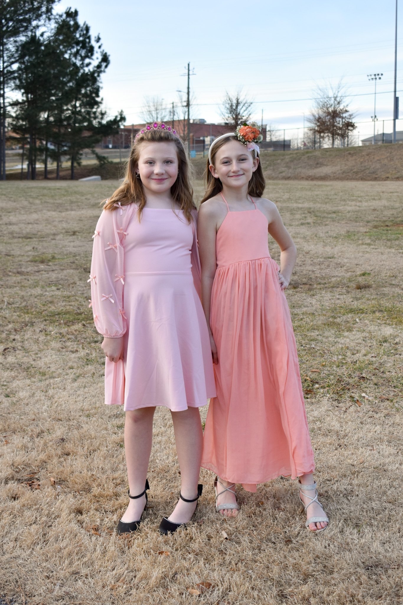 Two young girls dressed in pink dresses standing outdoors on a grassy field, holding hands, with trees and a sports field fence in the background.