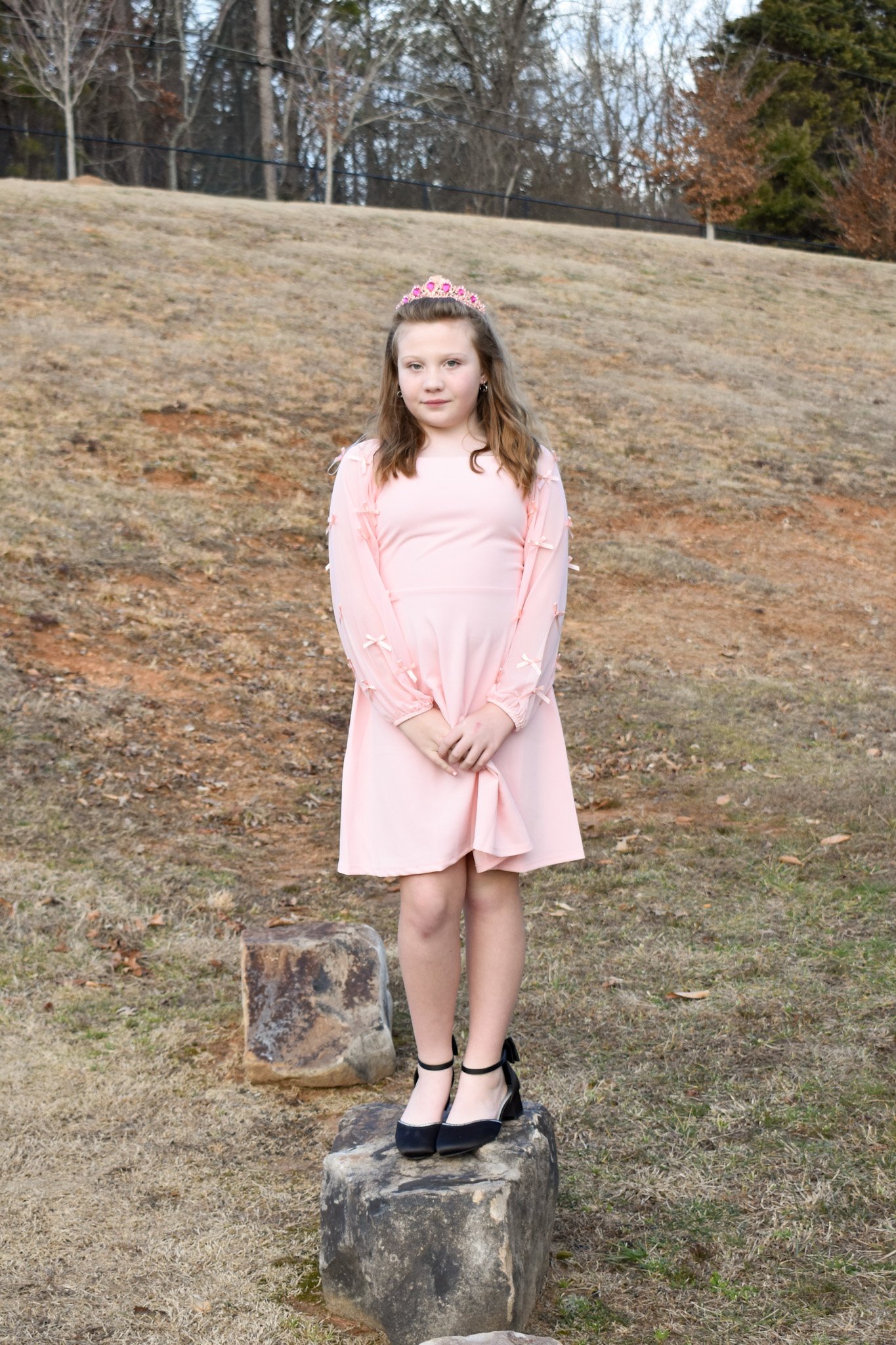 A young girl in a pink dress standing on rocks outdoors, wearing a small crown and black shoes.