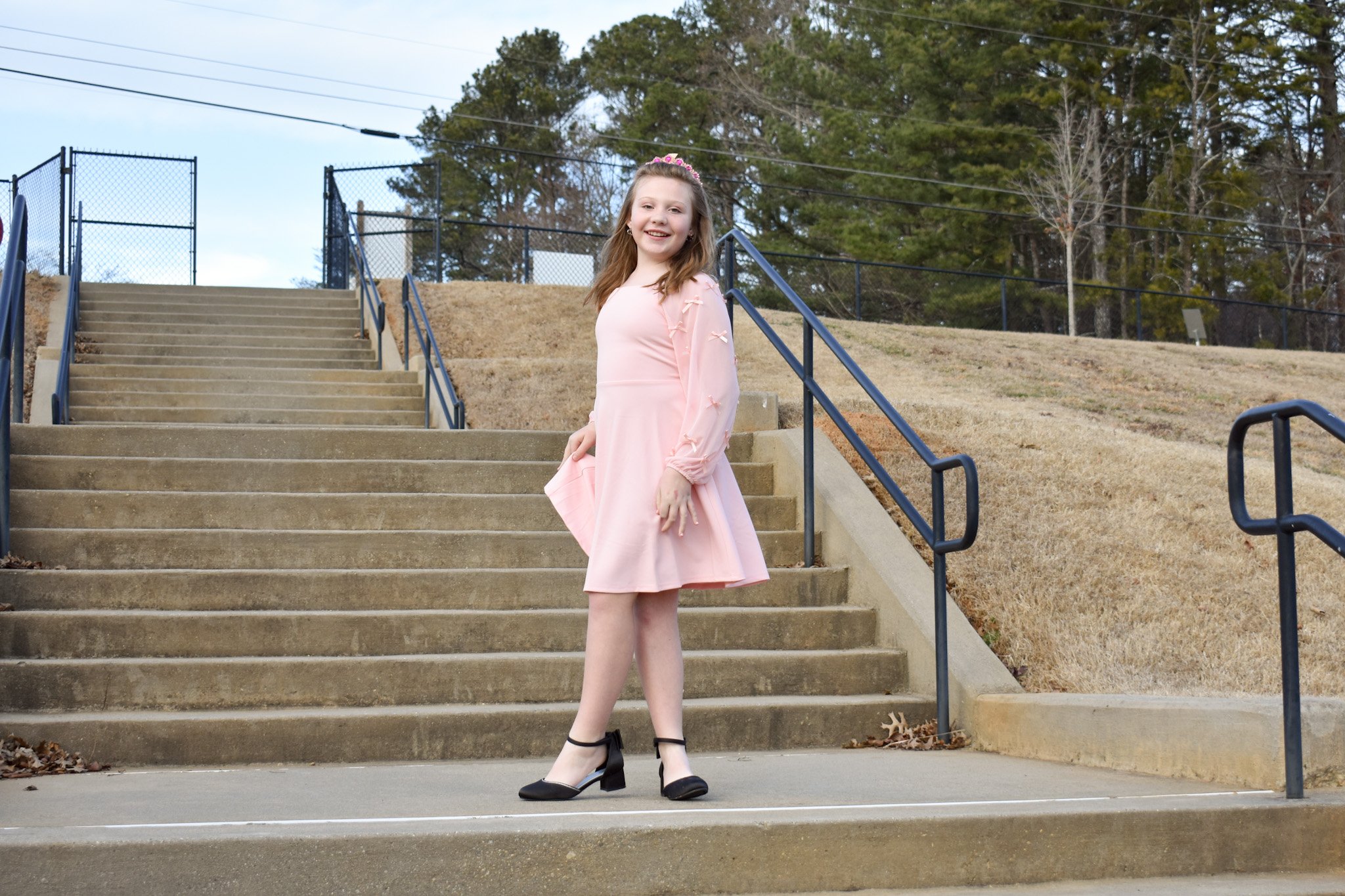 A girl in a pink dress and black heels stands on the steps outside, smiling at the camera.