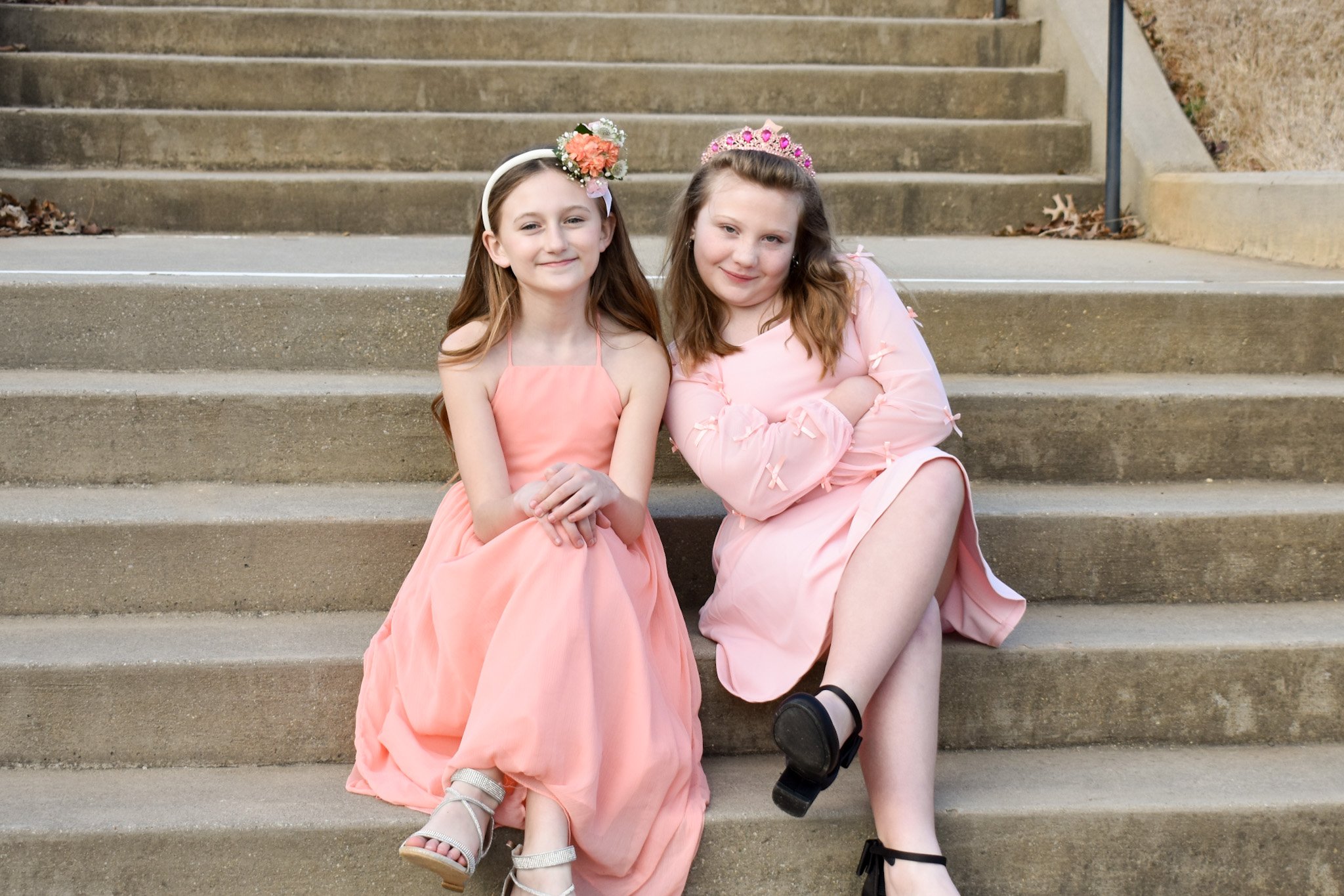 Two young girls wearing pink dresses sitting on outdoor concrete steps, smiling at the camera. One girl has a floral headband, and the other has a pink tiara.