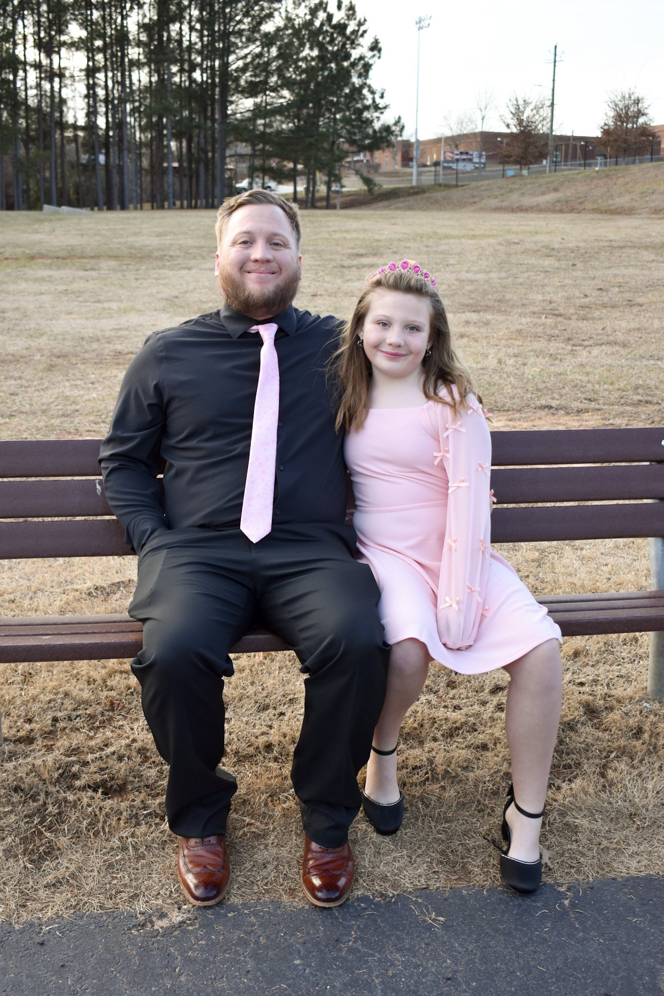 A man and a girl sitting on a park bench outdoors. The man is in a black shirt, pink tie, black pants, and brown shoes. The girl is in a light pink dress with long sleeves decorated with small bows, black shoes, and a pink tiara. They are smiling, wi