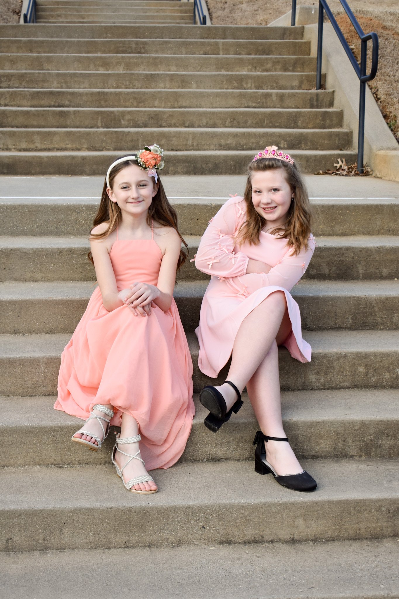 Two young girls sitting on outdoor concrete stairs, dressed in pink dresses with flower crowns, smiling at the camera.