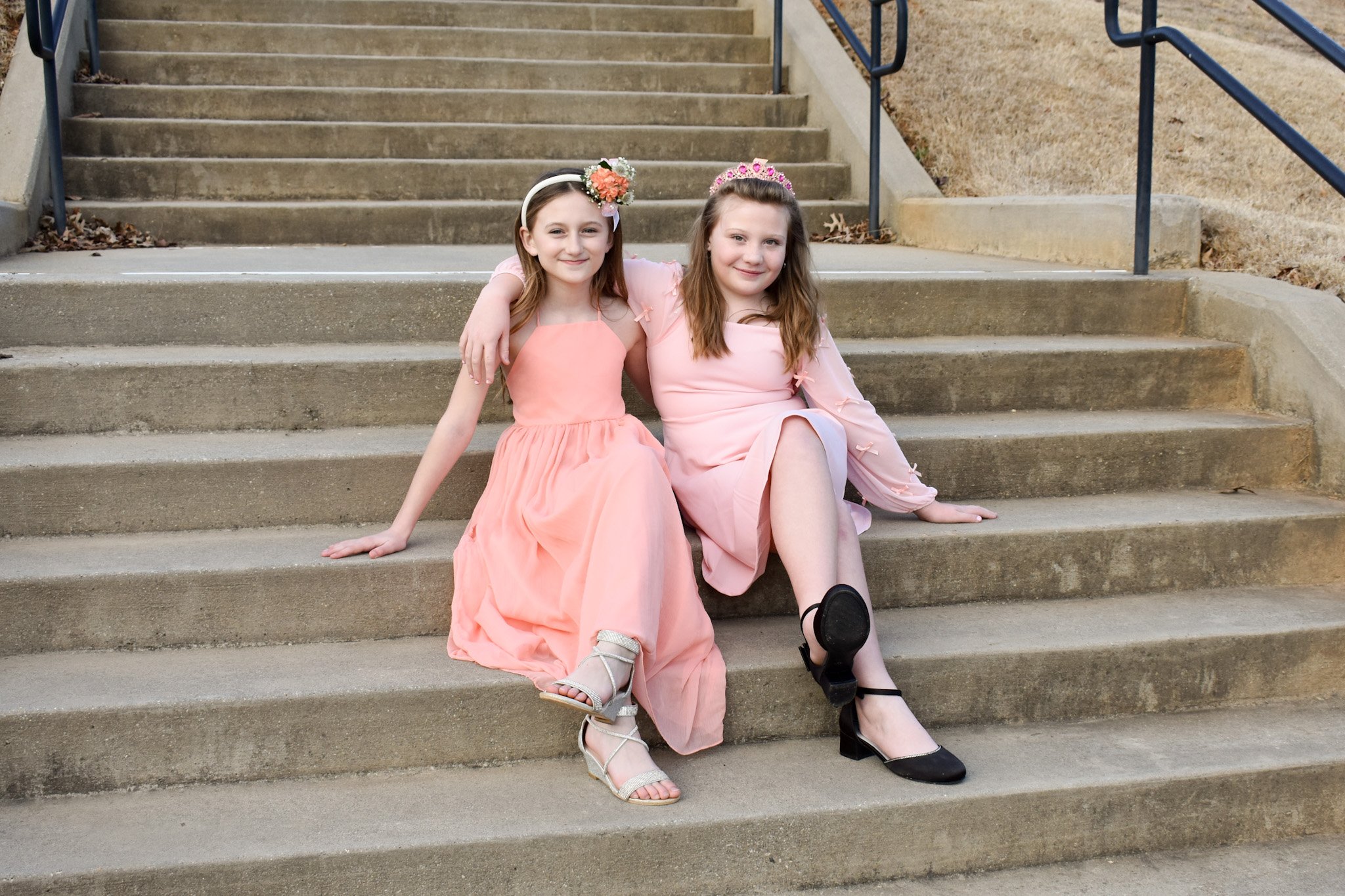 Two young girls sitting on outdoor concrete stairs, wearing pink dresses and floral headbands, smiling and embracing each other.