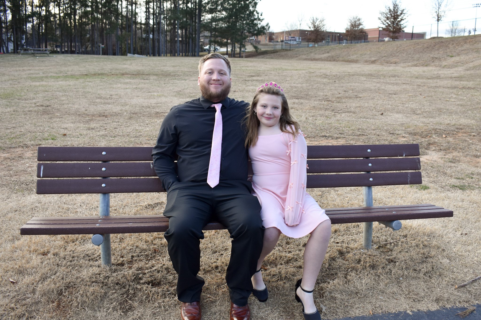 A man and girl sitting on a park bench. The man is wearing a black shirt, black pants, and a pink tie. The girl is wearing a pink dress and black shoes, with a pink headband. They are smiling and sitting close together outdoors.