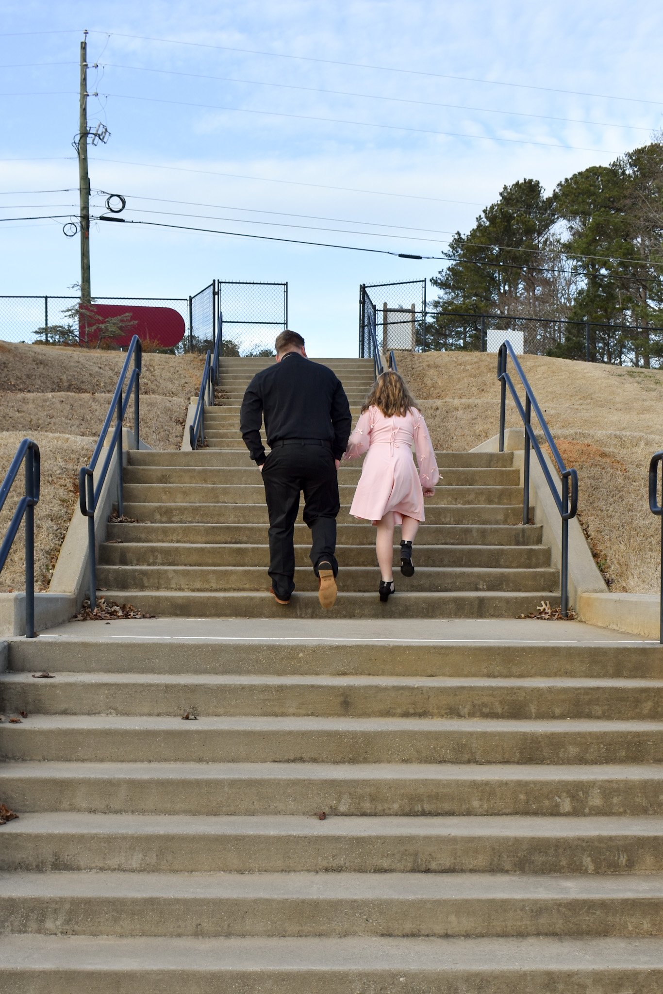 A man and a girl walking up concrete stairs outdoors, holding hands, with a school or park fence and trees in the background.