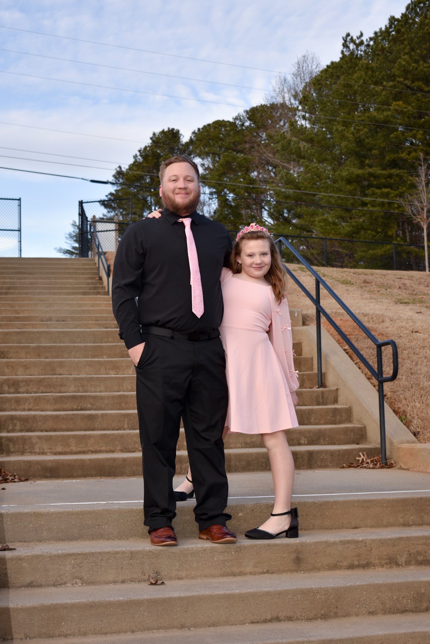 A man and a girl standing on outdoor stairs, smiling. The man wears a black shirt, pink tie, and black pants. The girl wears a pink dress and a tiara, with her arm around the man's waist.