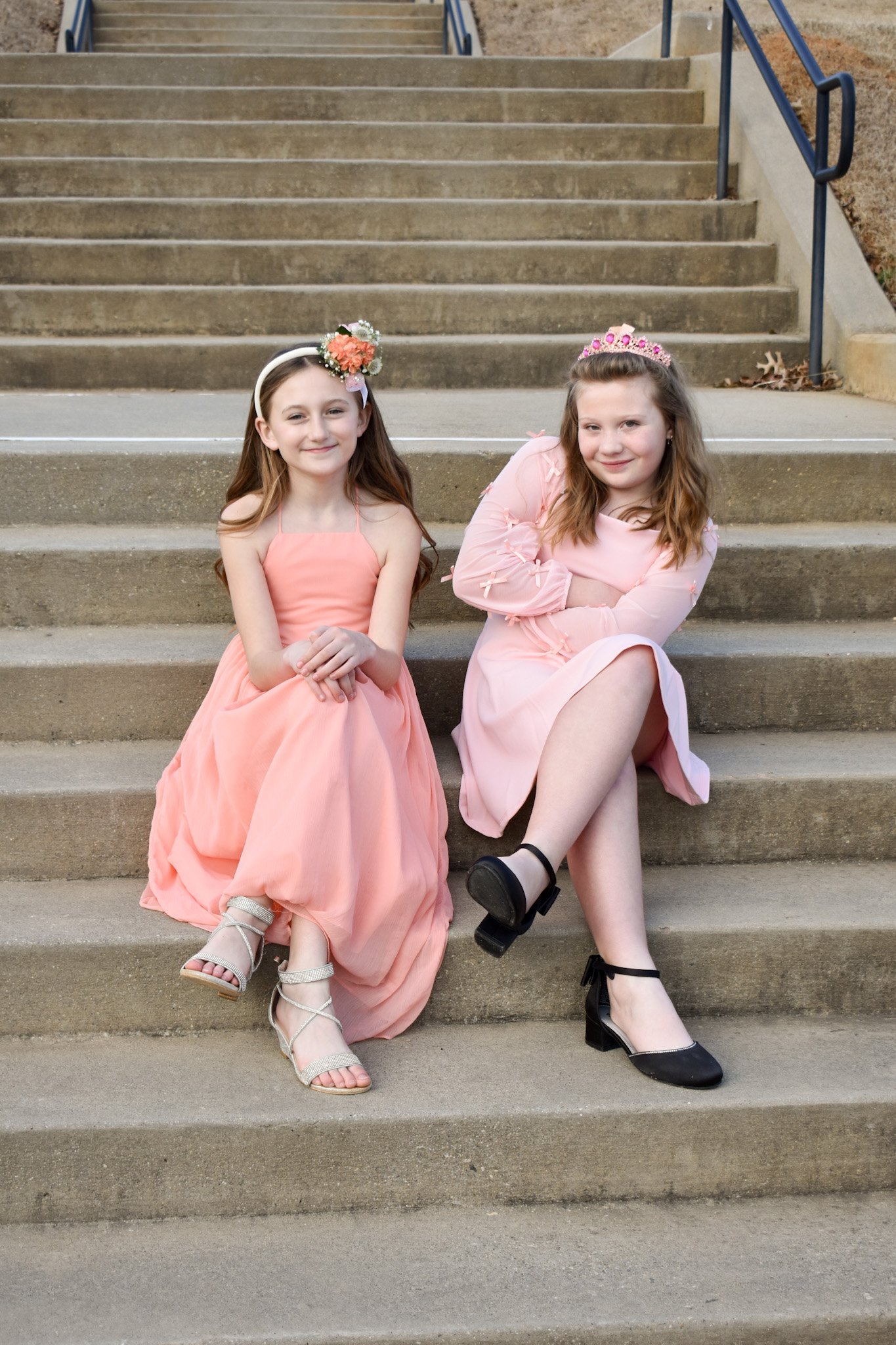 Two young girls sitting on outdoor concrete steps, dressed in pink formal dresses and wearing tiaras, smiling at the camera.