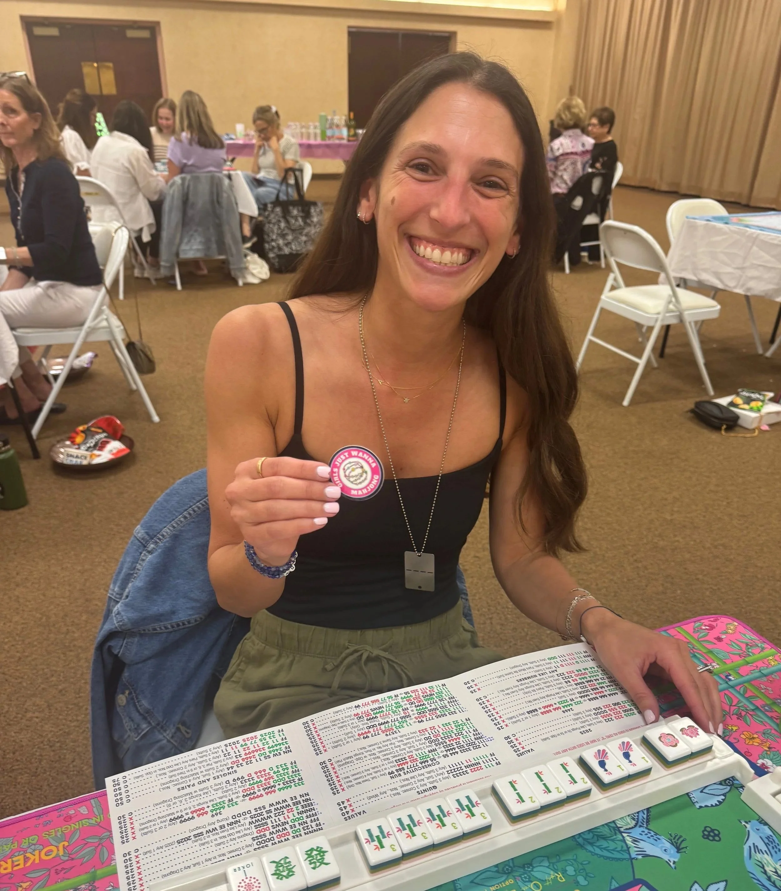 A woman smiling and holding a mahjong tile at a table with mahjong game pieces and score sheets, in a lively indoor setting with other people in the background.