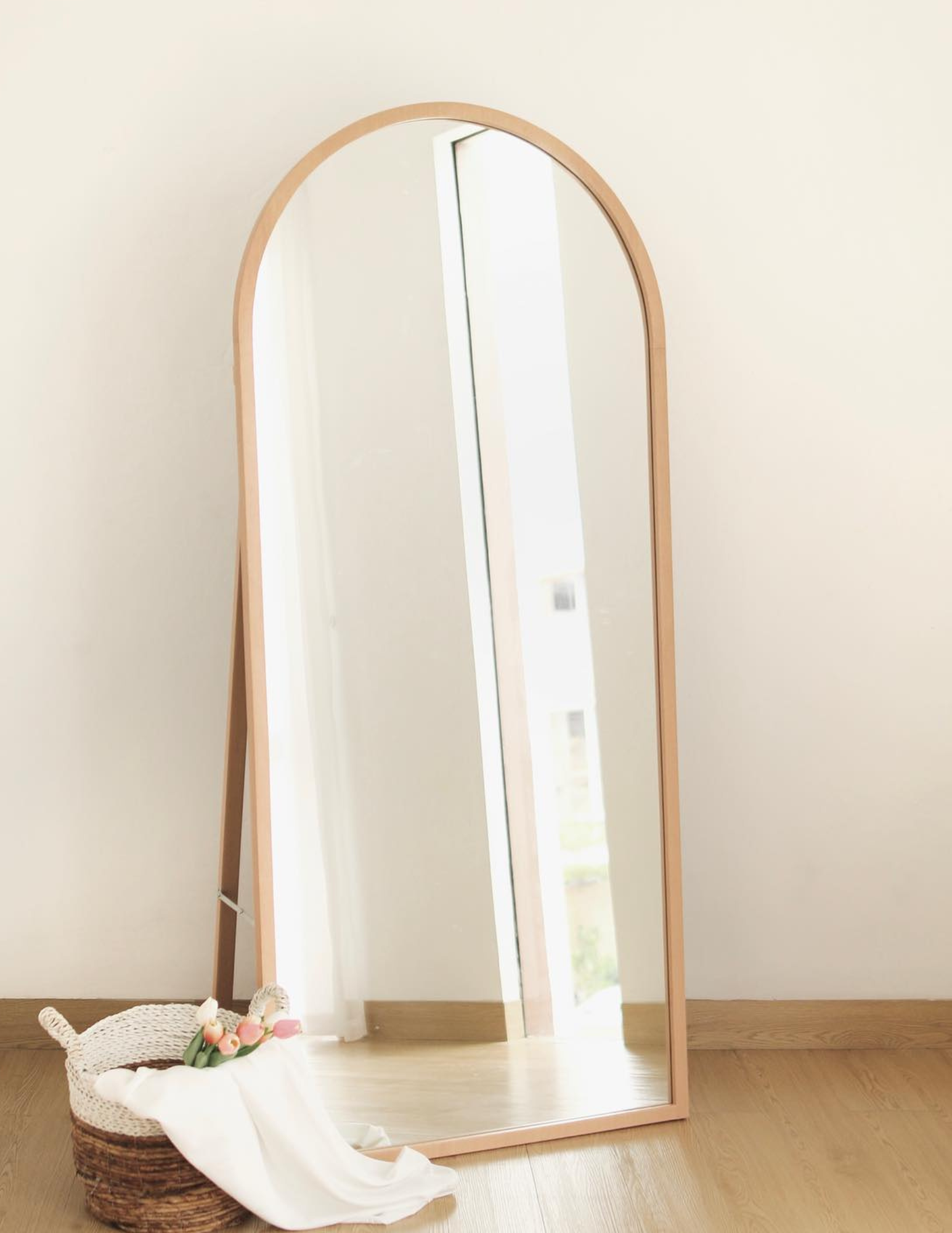 Full-length arched mirror with wooden frame leaning against a white wall on a wooden floor, next to a woven basket holding a white cloth and tulips.