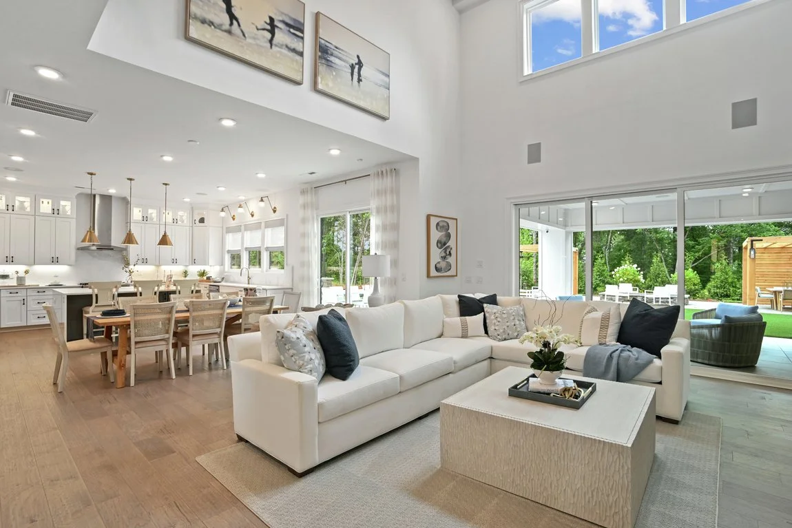 Modern open-concept living room with white sectional sofa, decorative pillows, and a coffee table. The room features large windows with a view of the outdoor area, and a kitchen with a wooden dining table and white cabinetry. The space has high ceilings and contemporary décor.