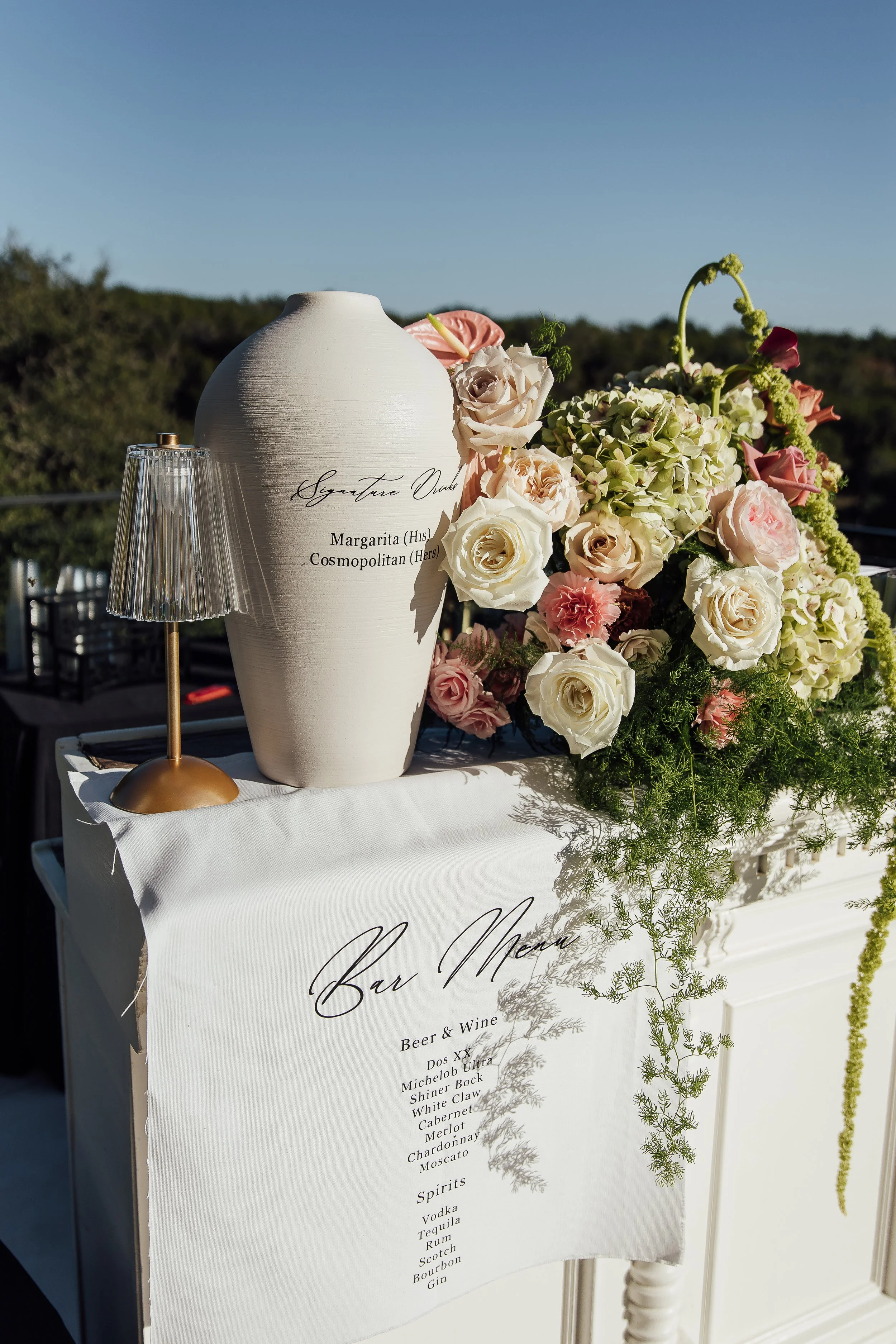 A wedding bar menu displayed on a white cloth-covered table, decorated with a bouquet of white and pink roses and greenery, and a white vase with text.