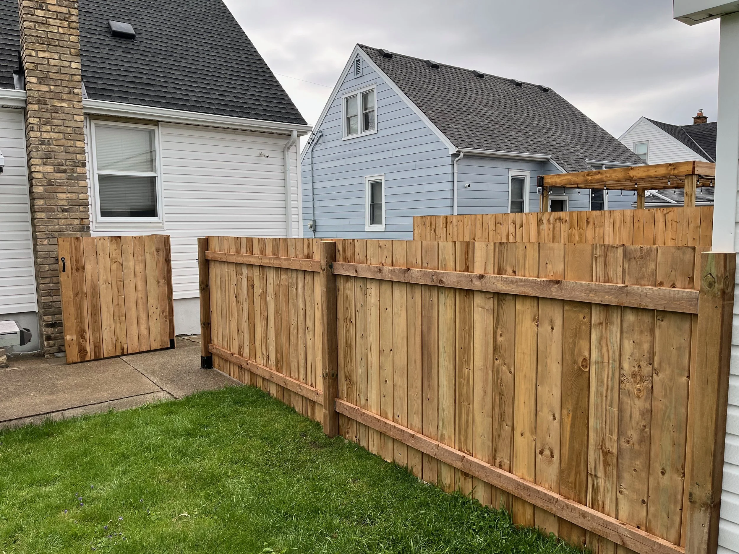 New wooden privacy fence installed in backyard, with one gate leaning against the house. Green grass and neighboring houses visible.