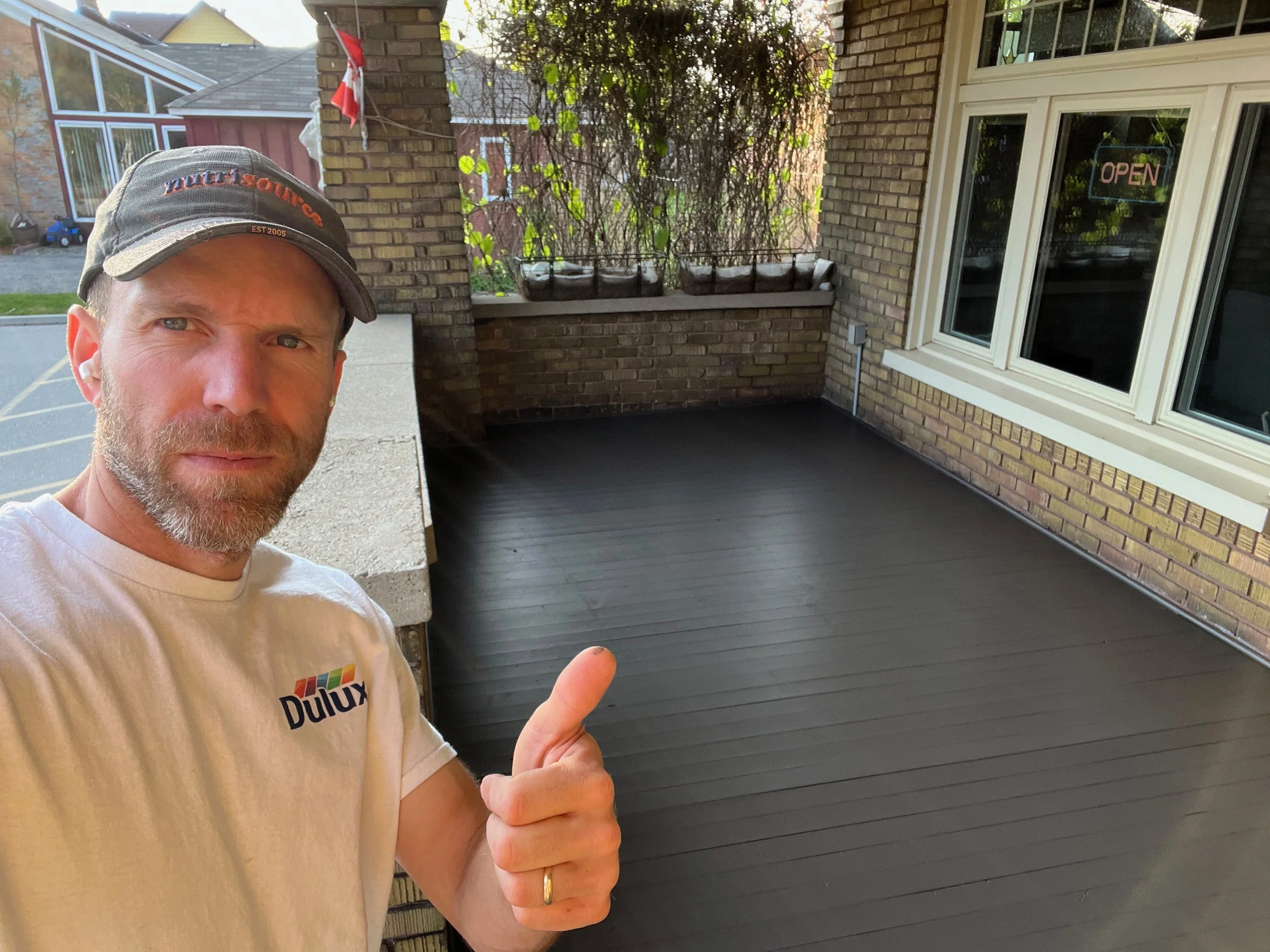 Man in a white Dulux shirt and gray cap taking a selfie on a newly painted black deck outside a brick house, giving a thumbs-up.