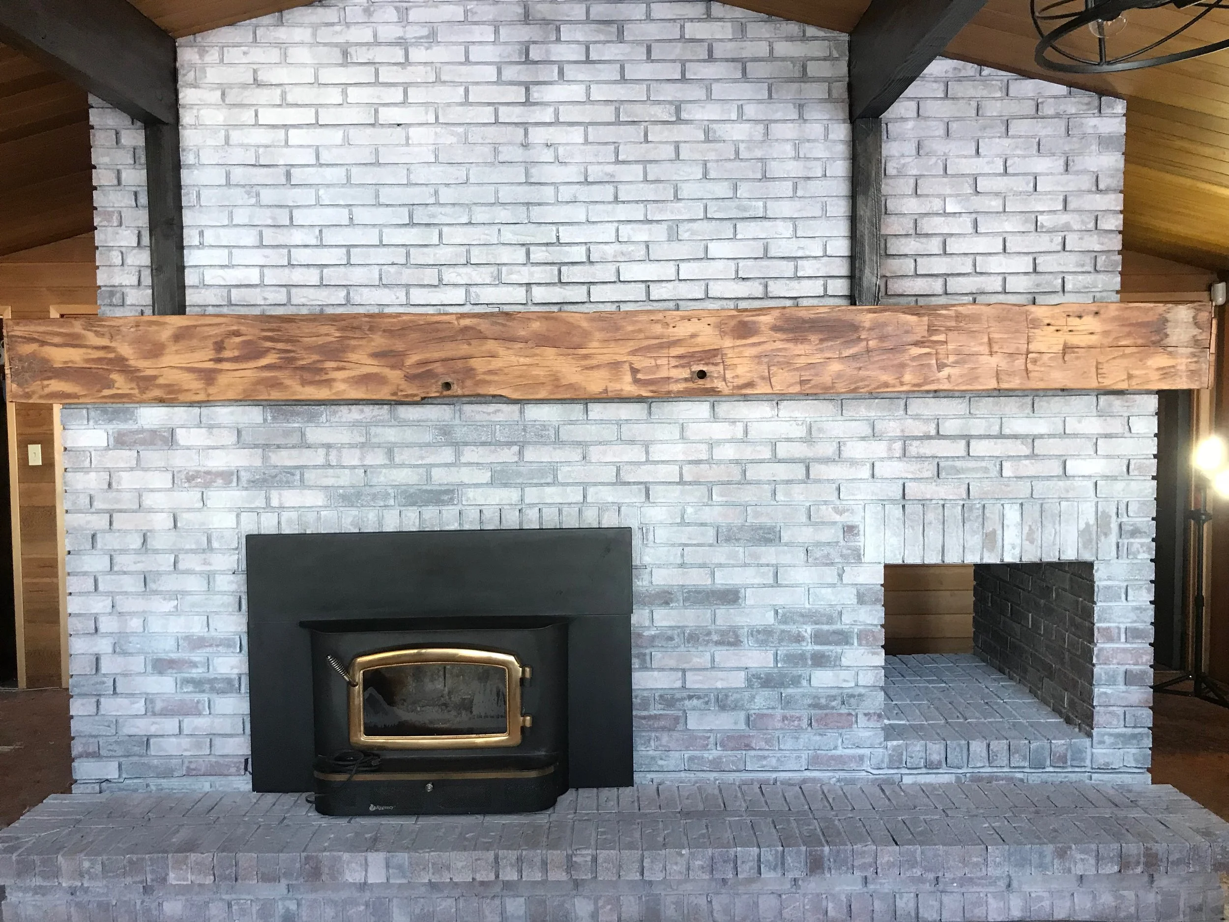 Brick fireplace that has been white washed with a wood mantel and a wood stove insert, set in a living room with wooden walls and ceiling.