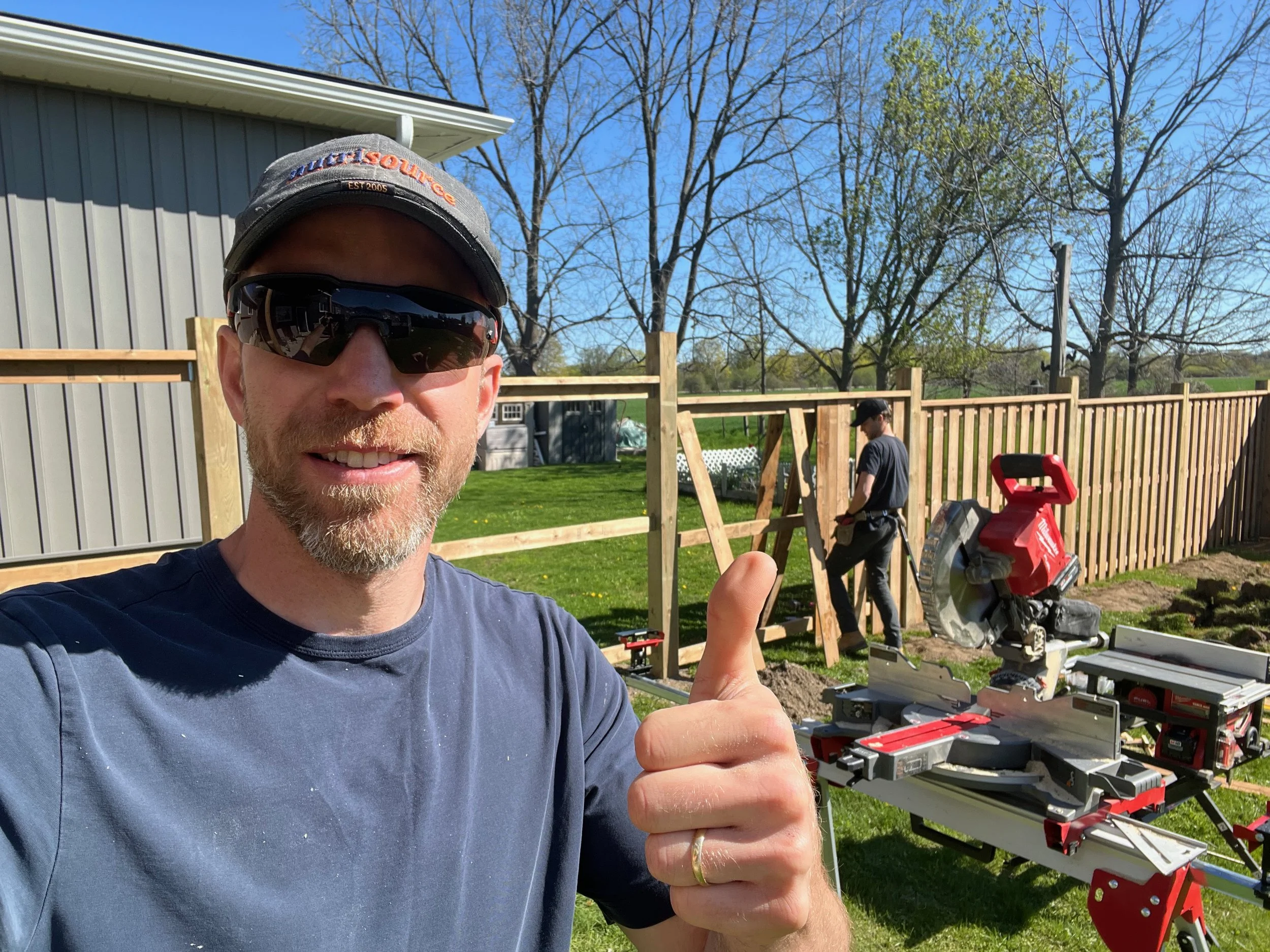 A man wearing sunglasses and a cap takes a selfie with a smile and a thumbs-up gesture, standing outdoors during daytime. In the background, another person is working on a wooden fence near a miter saw, with a green lawn, trees, and a gray building visible.