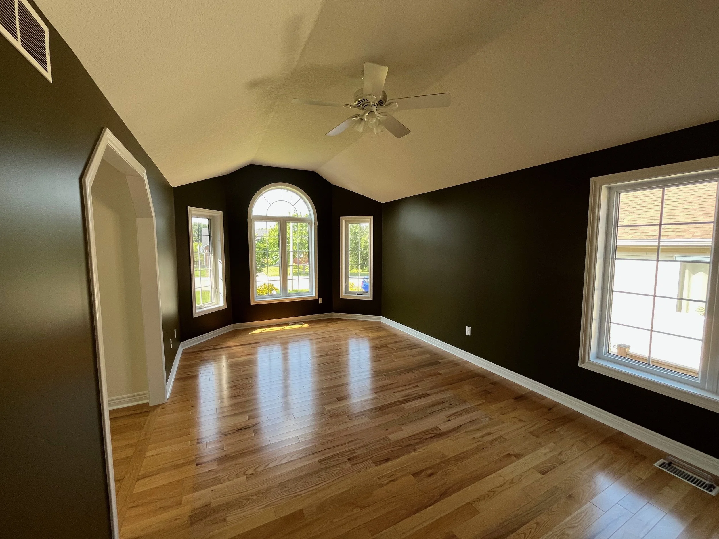 Empty living room with hardwood floors, dark accent walls, three windows, and a ceiling fan.