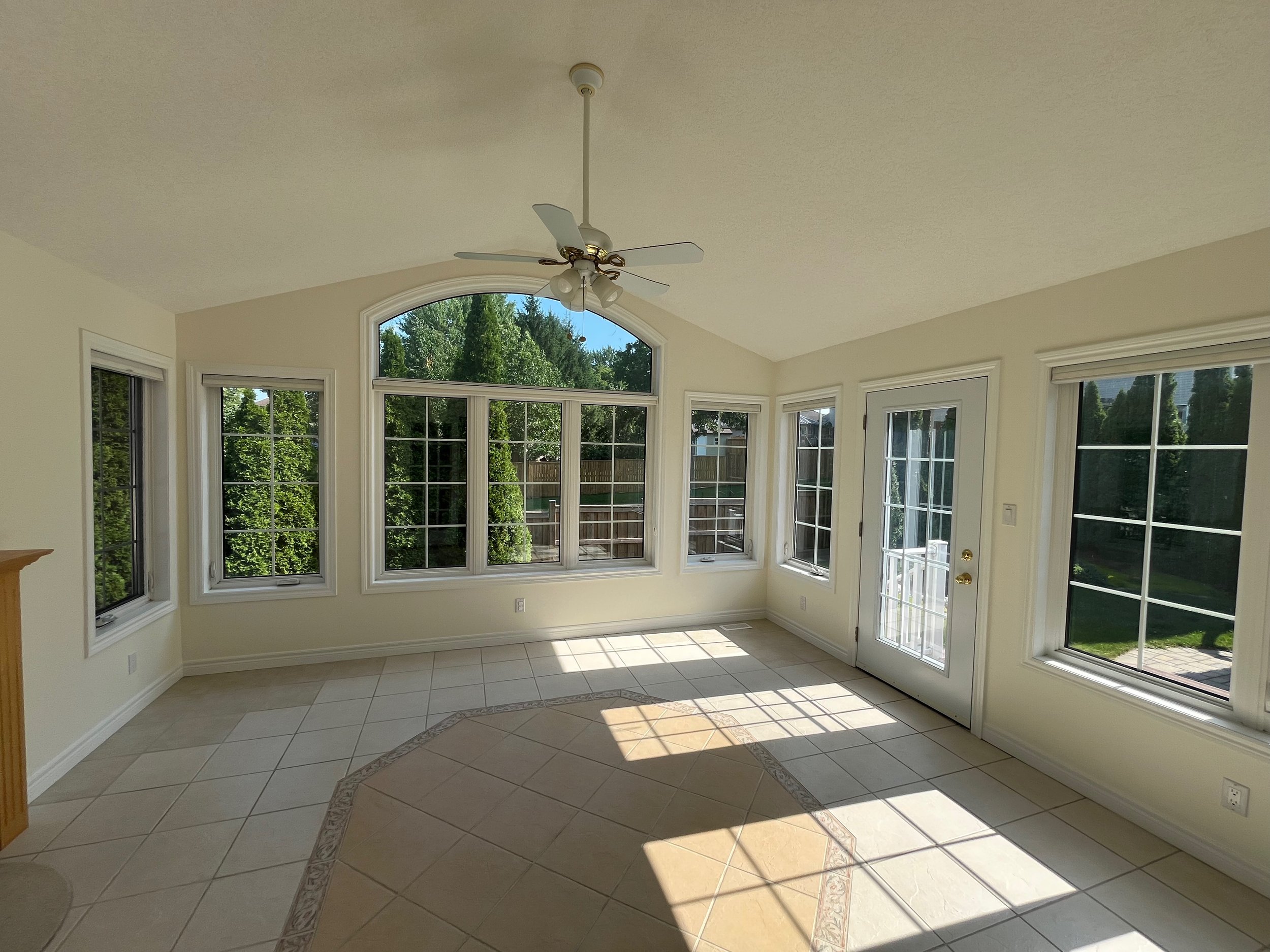 Empty sunroom with large windows, a ceiling fan, a tiled floor, and a door leading outside, with sunlight streaming in.