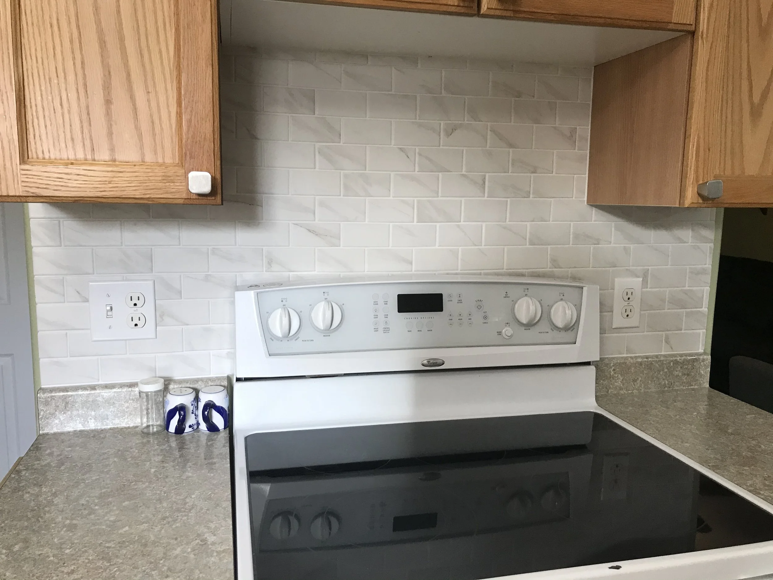 A kitchen countertop with a white electric stove, two white cabinets above, a backsplash with white subway tiles, and two small containers near an electrical outlet.