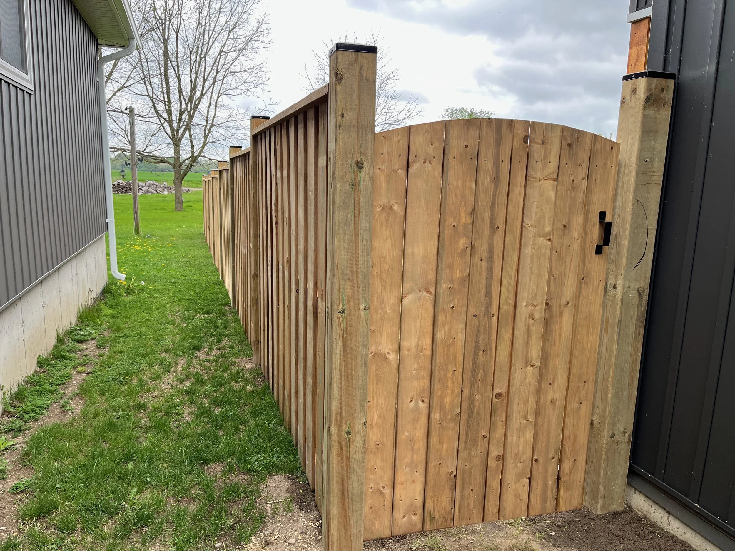 A wooden privacy fence with a gate runs alongside a gray building and a grassy yard with a tree and cloudy sky in the background.