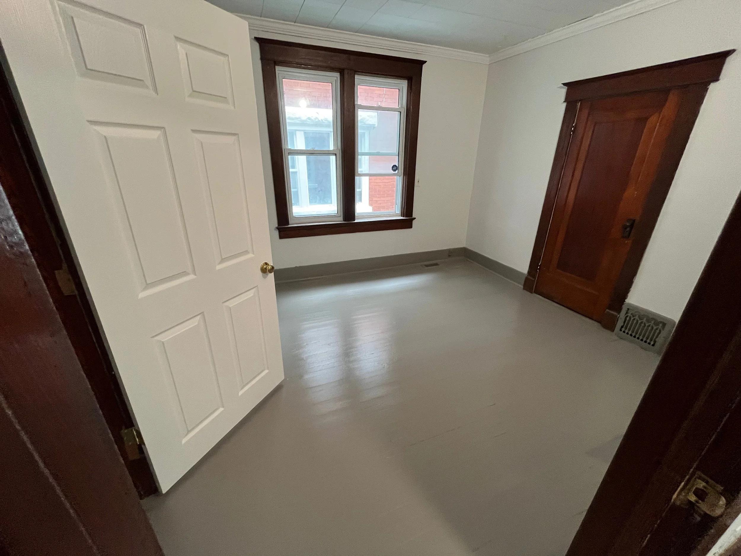 An empty room with white walls, a freshly painted floor, a window with dark wood trim, a closet with a wooden door, and a vent near the floor.