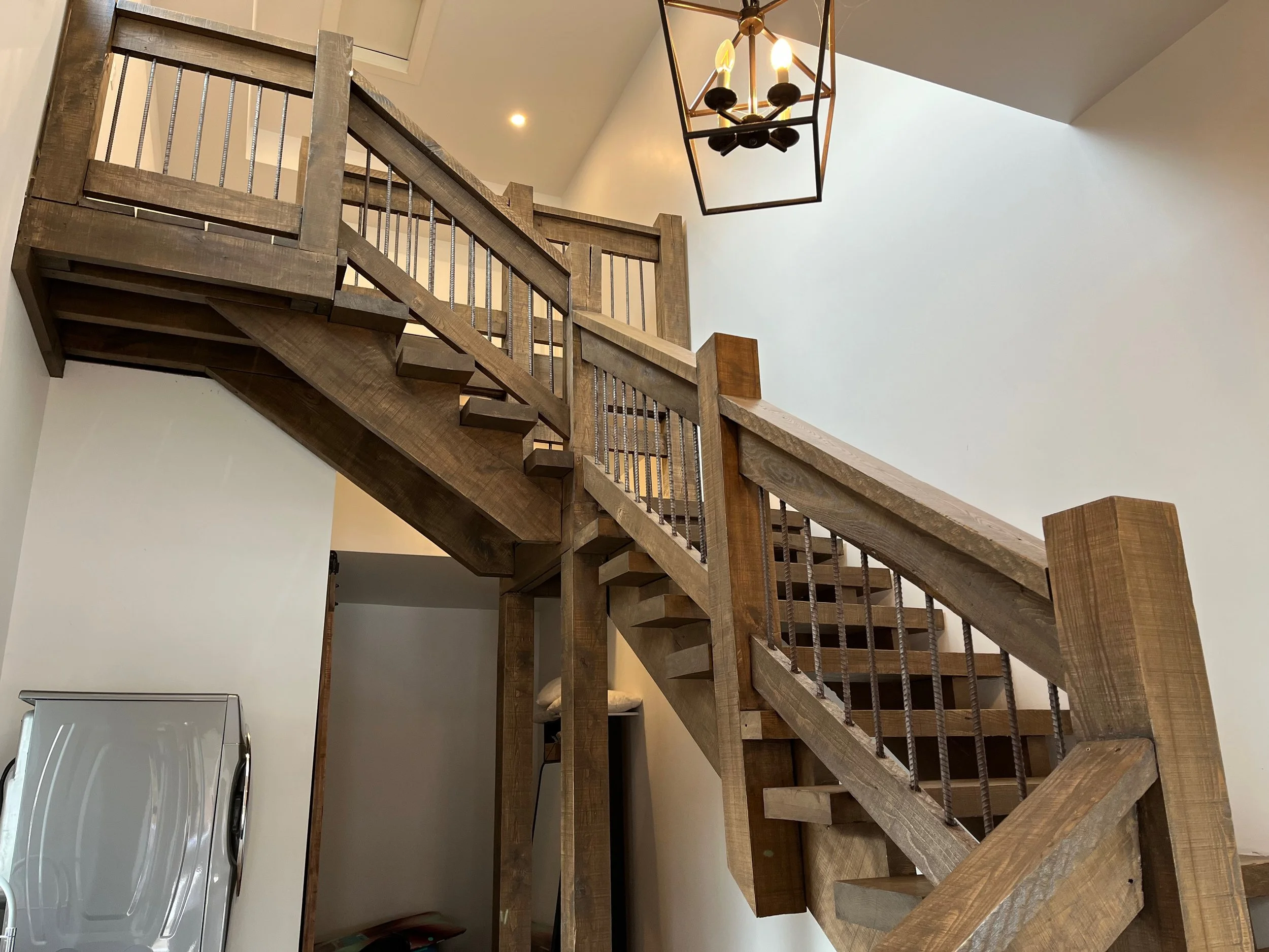 Wooden staircase, freshly stained and sealed, leading up to an upper level with a modern chandelier hanging from the ceiling.