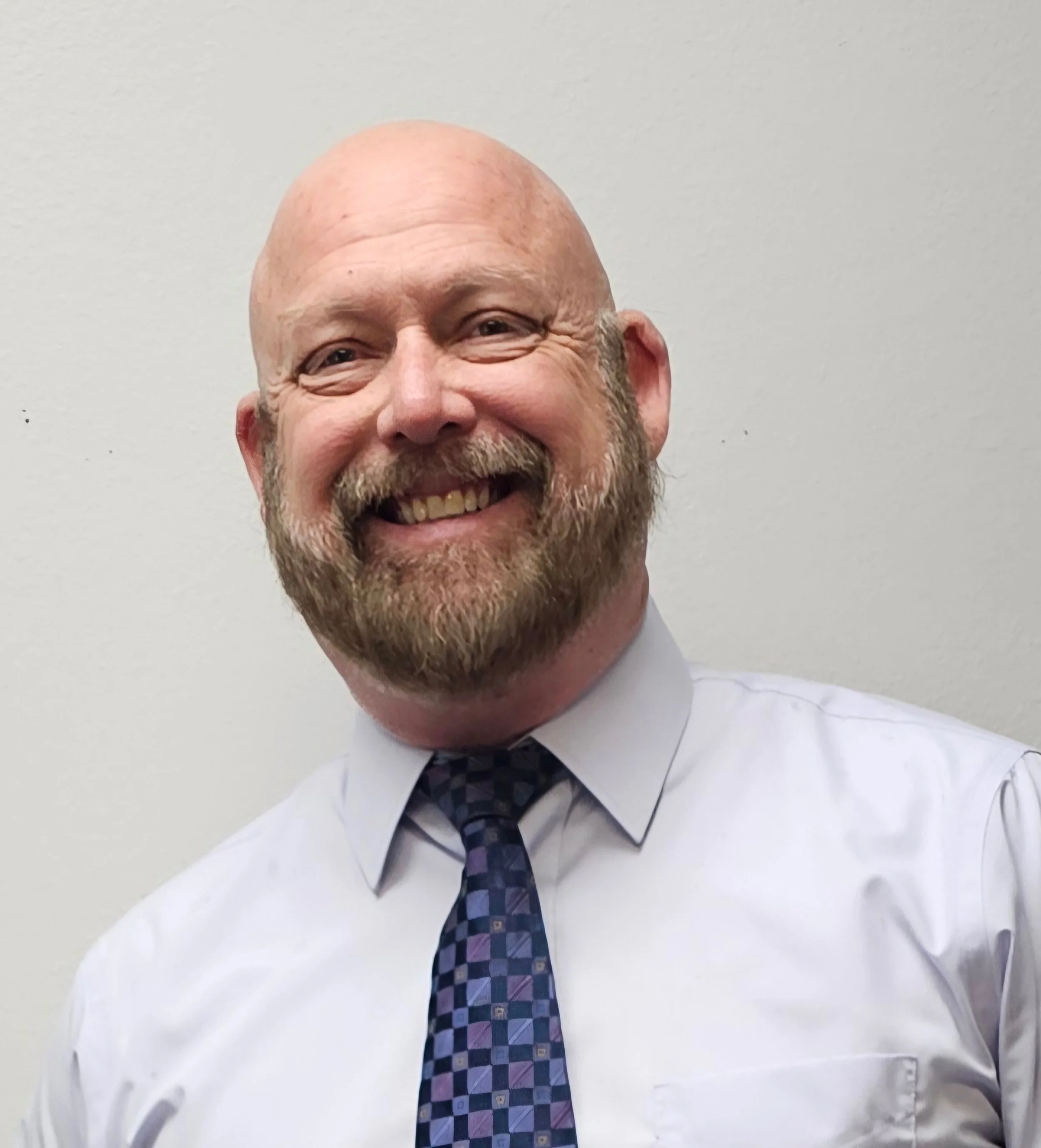 A smiling man with a beard and bald head, wearing a white shirt and a striped tie, standing against a plain wall.