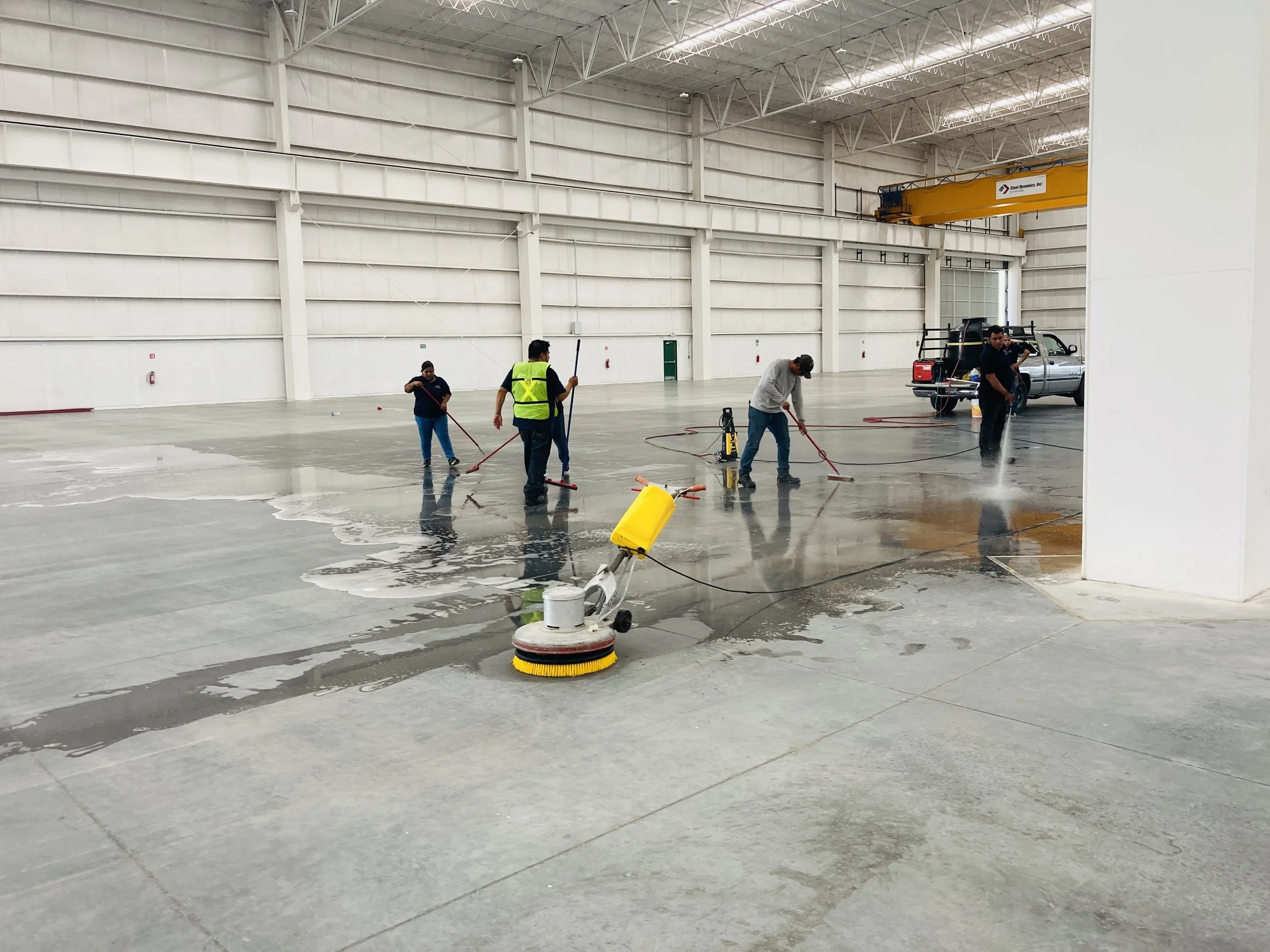 Personal de limpieza trabajando en el pulido y limpieza del piso en un interior de un almacén o hangar grande con techo alto y estructura metálica.