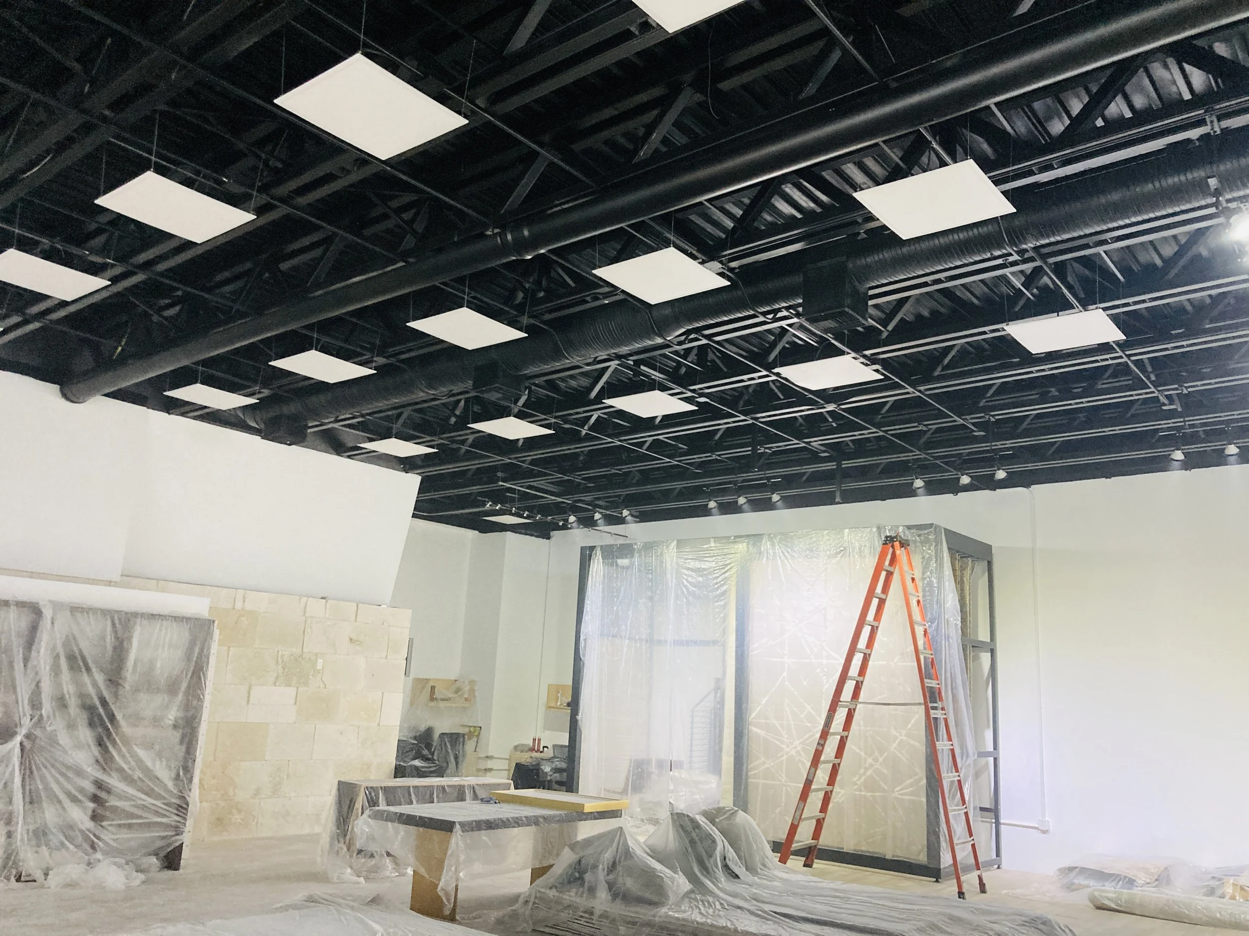 Sala en construcción con techo expuesto de tuberías negras y paneles de cielo falso, andamios cubiertos con plástico, una escalera roja y diversas herramientas y materiales de construcción.