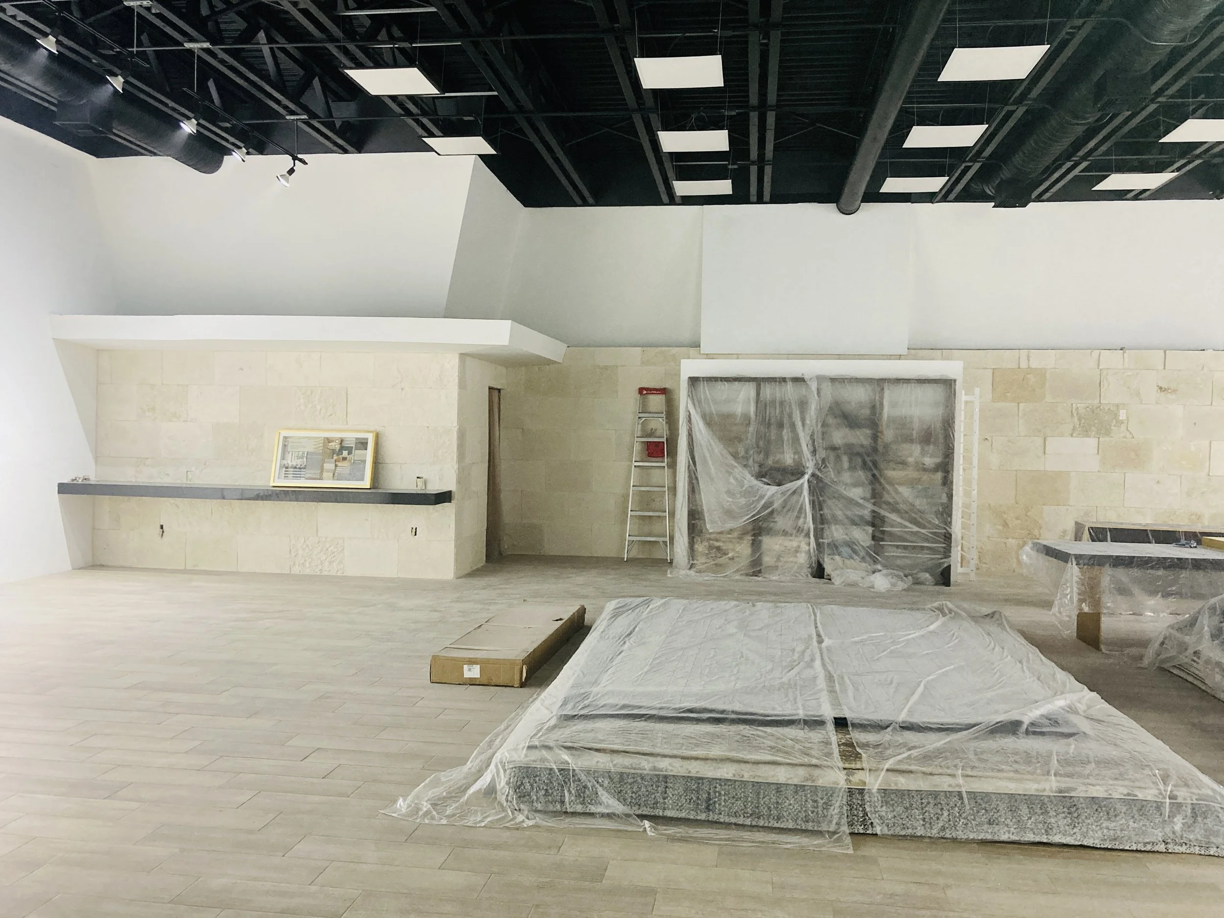 Interior de un espacio en construcción, con paredes de piedra clara, piso de madera clara, y equipo de construcción cubierto con plástico, entre ellos una estructura grande en la esquina derecha y una escalera roja apoyada en la pared del fondo.