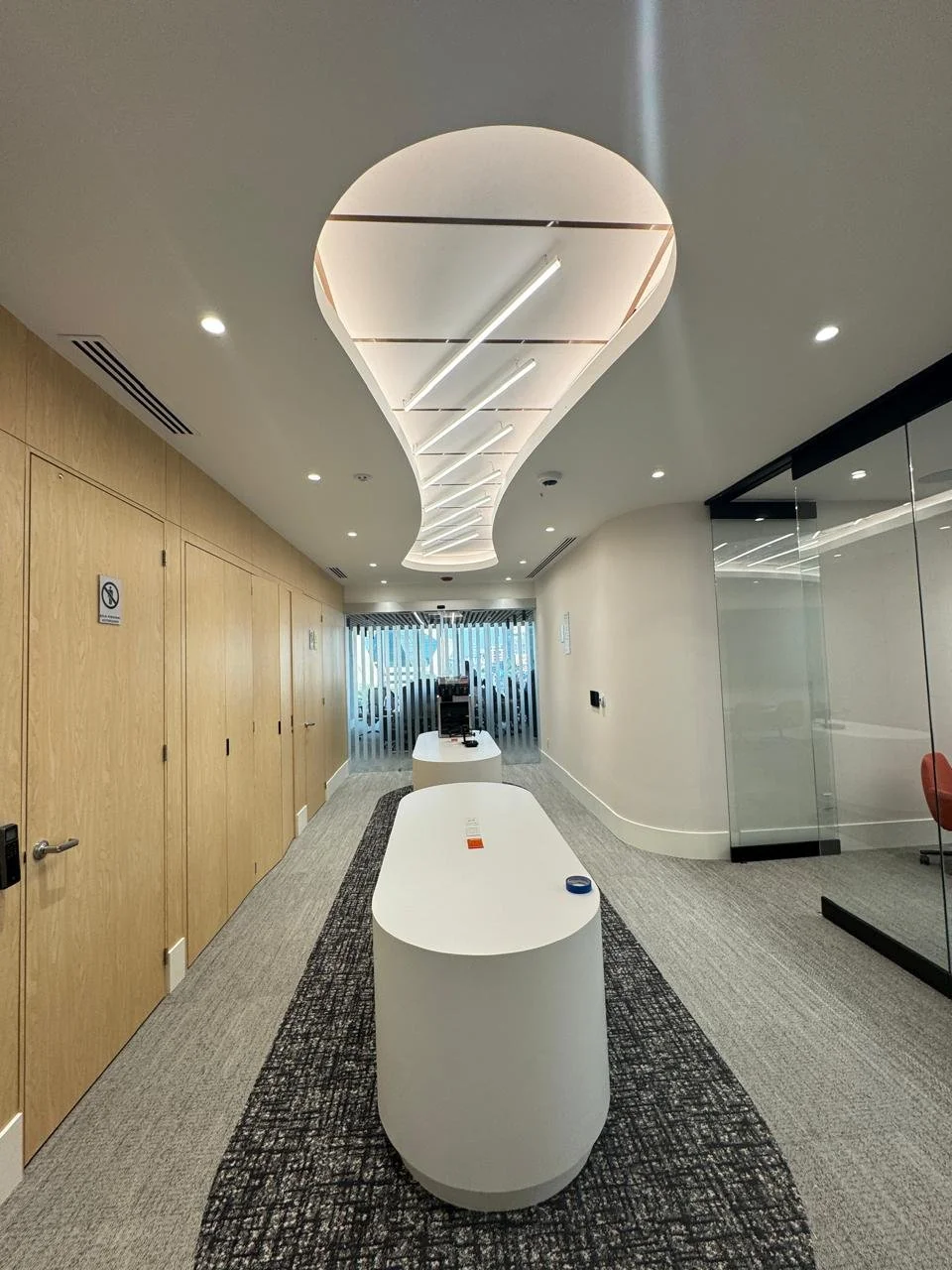 Interior moderno de oficina con mesas blancas y áreas de vidrio, iluminación LED en el techo y paredes de madera y cristal.