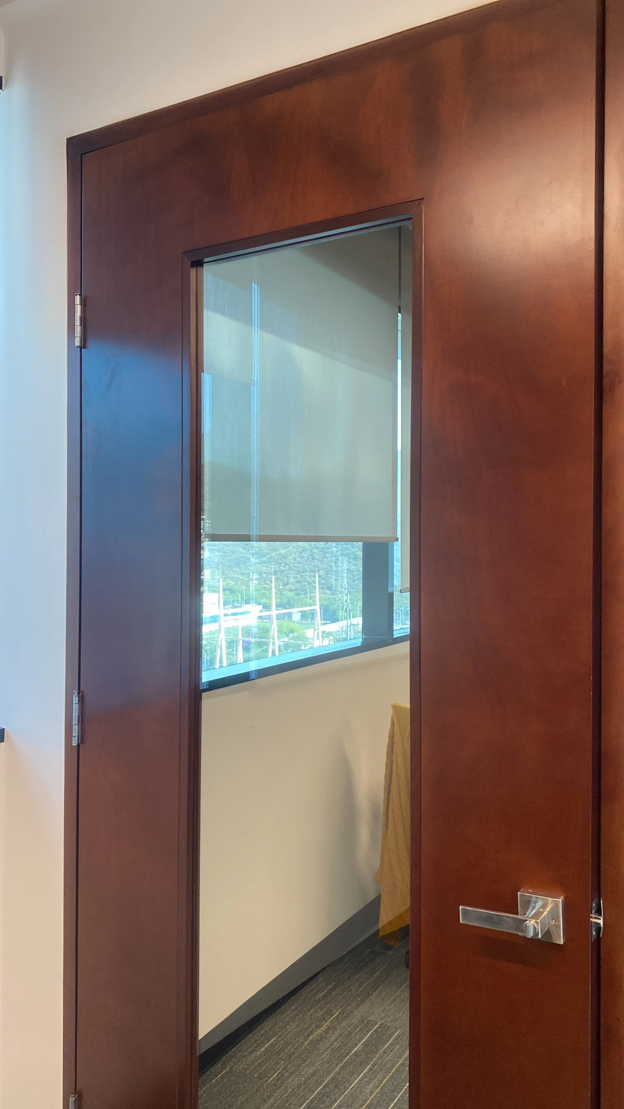 Puerta de madera marrón que da a una oficina con una ventana que tiene una persiana half opaca y una vista del exterior.