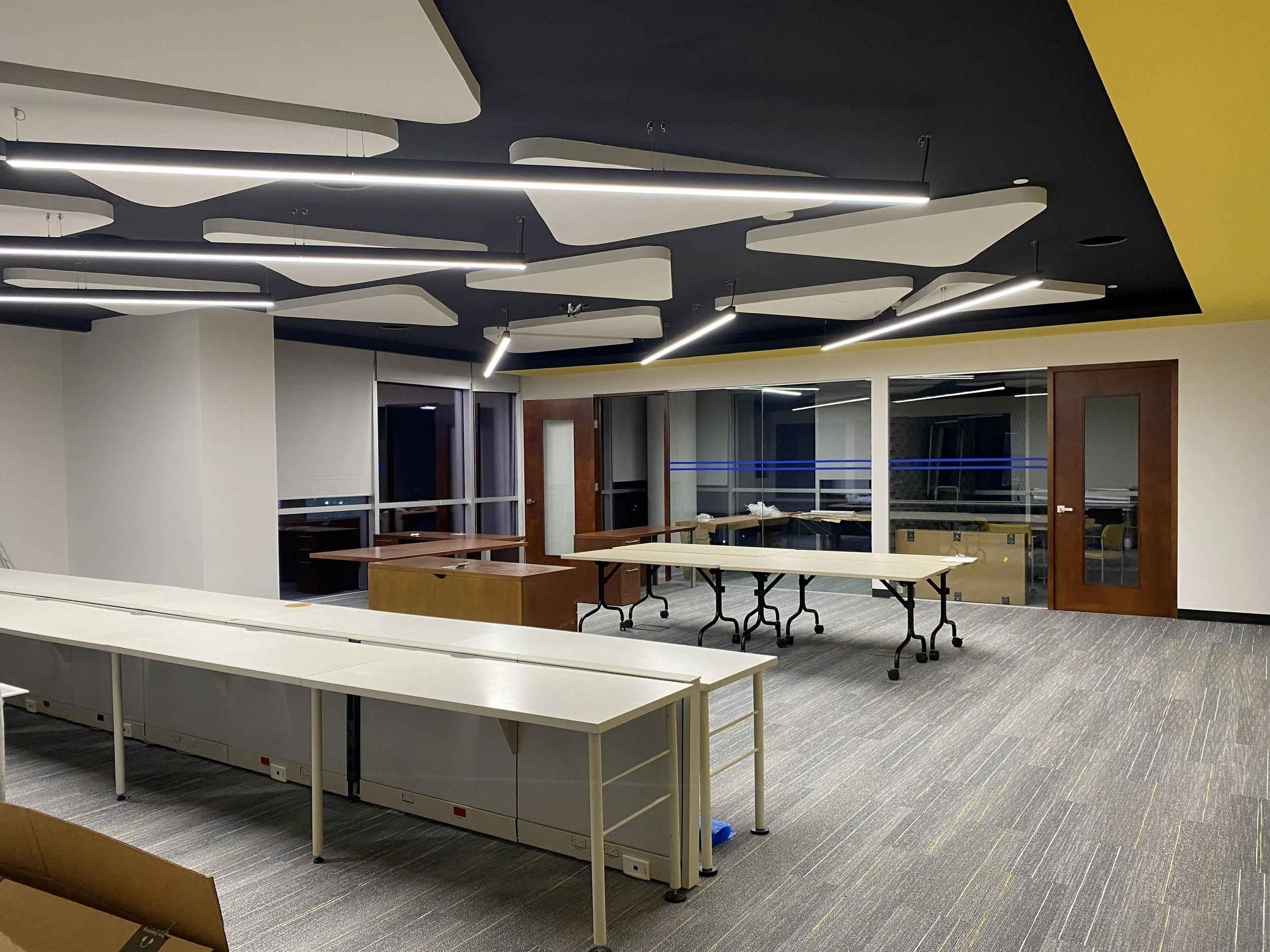 Sala de reuniones o oficina moderna con paredes blancas, ventanas grandes con vista al exterior, mesas de trabajo blancas y en tonos madera, lámparas delgadas y lineales en el techo negro decorado con formas blancas y amarillo en el techo, y piso de alfombra en tonos grises.