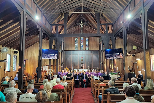 A church interior with wooden ceiling and walls, congregation seated in pews, and a choir or group of people standing in front of the altar.